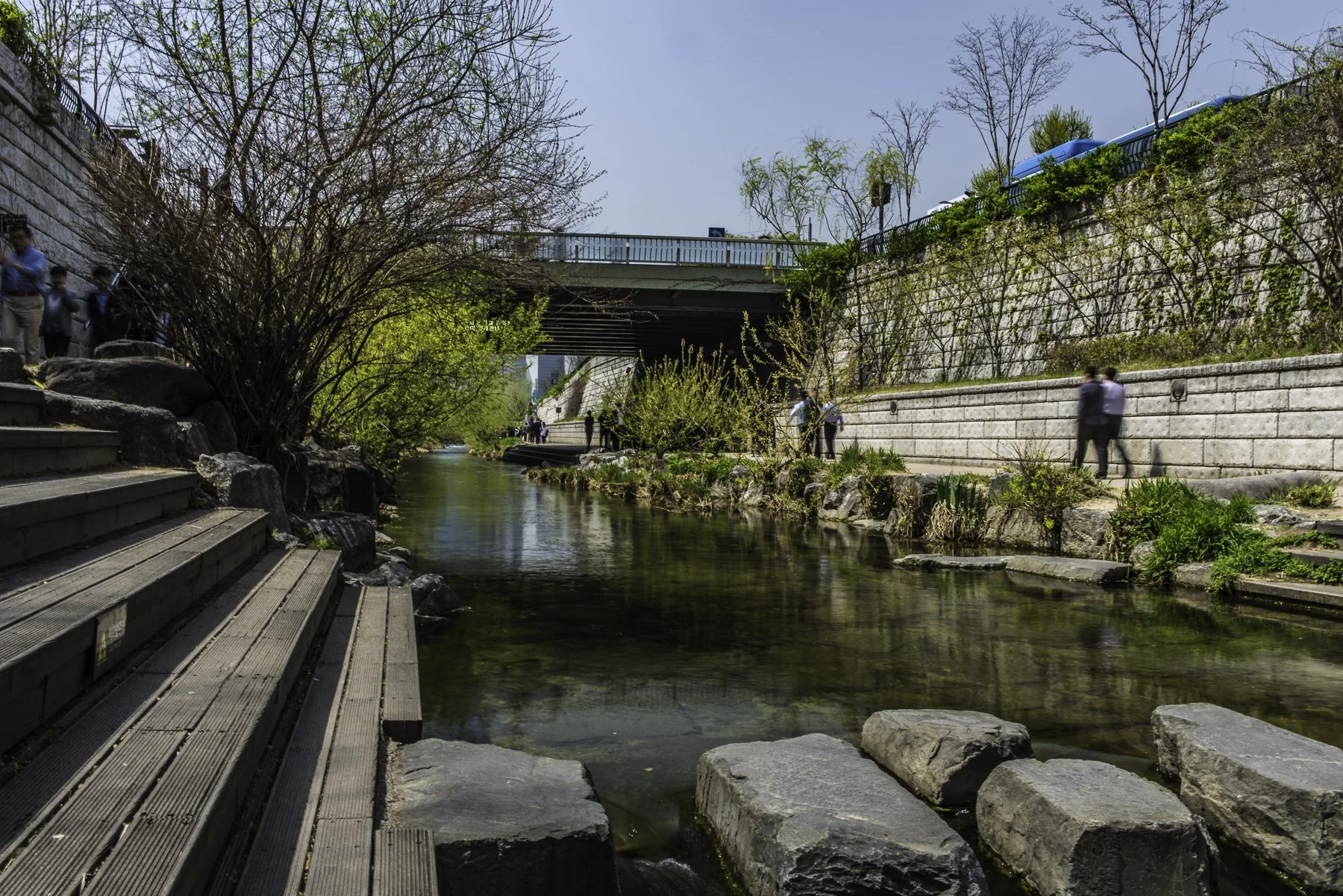 Urban park with a creek running through it, surrounded by rocks and greenery, with people walking along the pathways under a bridge on a sunny day.
