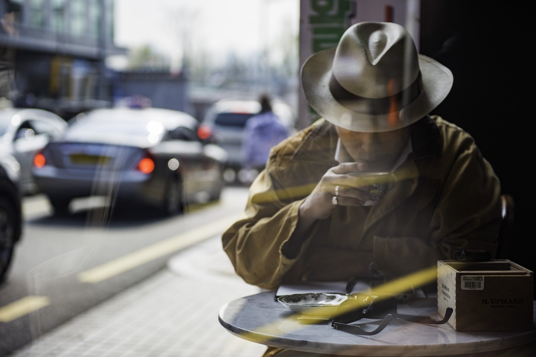 A person wearing a beige jacket and a beige hat sitting inside a cafe, looking at their phone. The photo is taken through a window, with blurred cars and pedestrians outside.