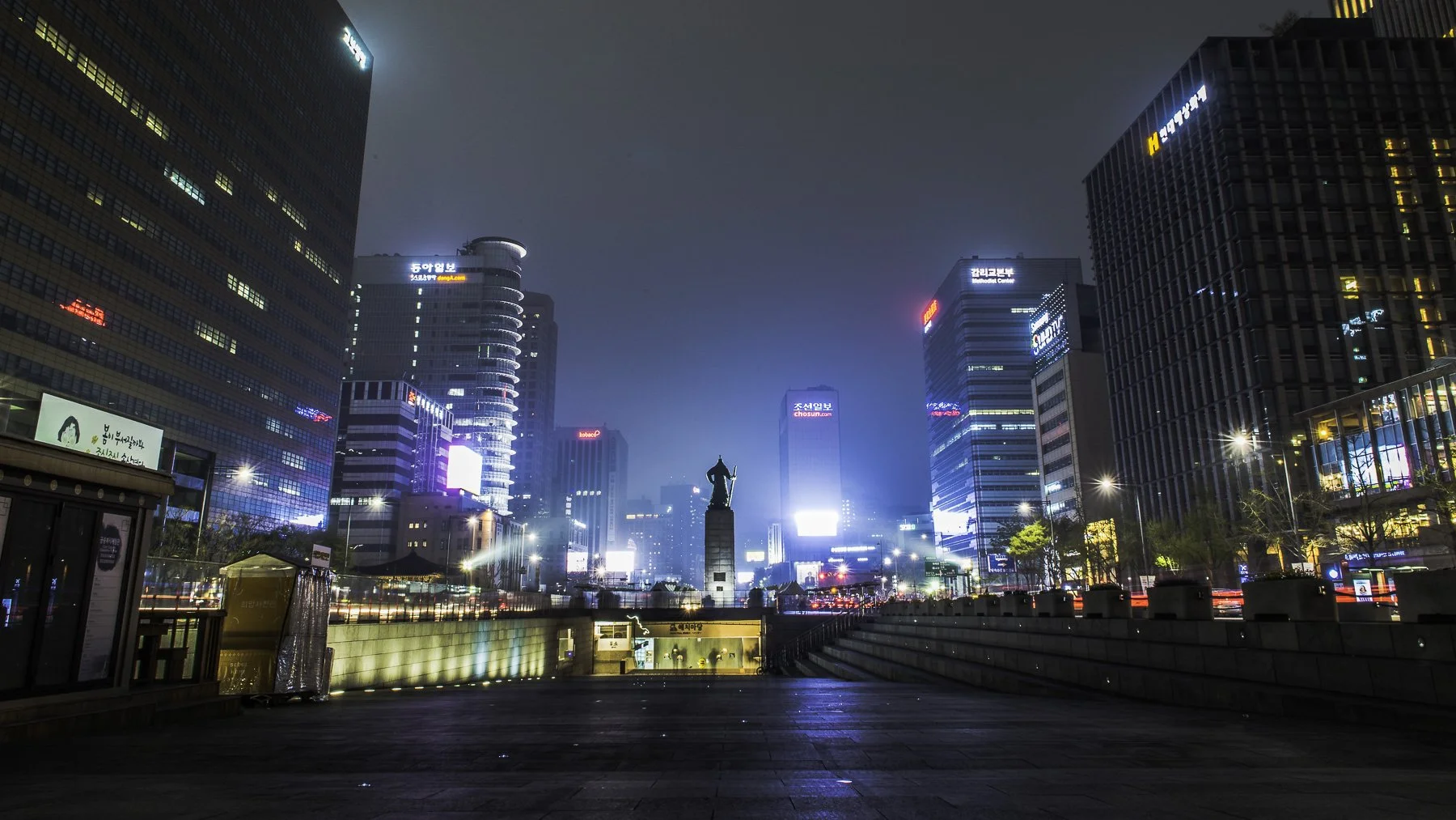 Night view of a city square with tall illuminated buildings and a statue in the center, with light trails from moving vehicles.
