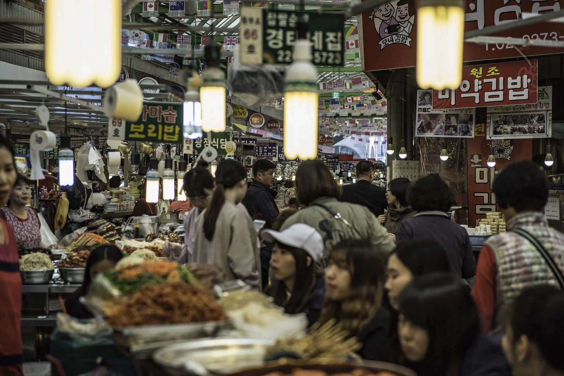 Crowded indoor market with various food stalls, vendors, and shoppers. Decorated with international flags hanging from the ceiling, including South Korea, Canada, and the United Kingdom. Bright lighting and signage in Korean advertise different food 