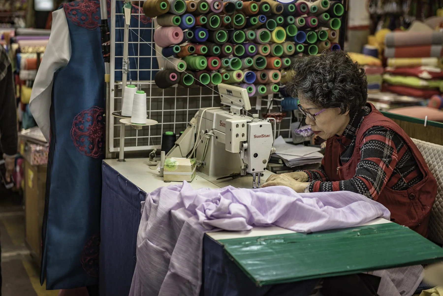 A woman working at a sewing machine in a fabric store with rolls of colorful fabric and sewing supplies around her.