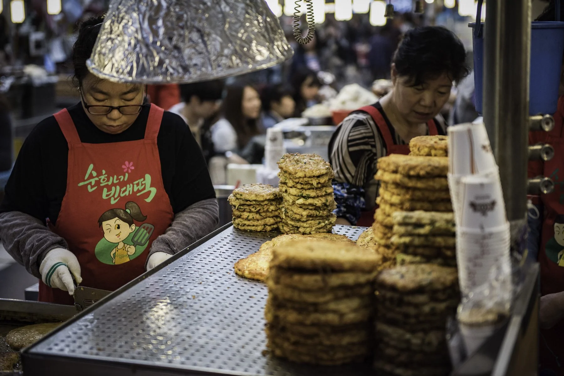 Street food vendor selling various fried snacks at an outdoor market, with two women in red aprons preparing and handling food, and a crowd in the background.