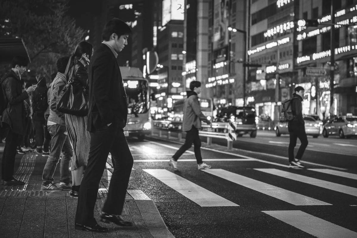 People crossing a city street at night, with illuminated signs and traffic around them.