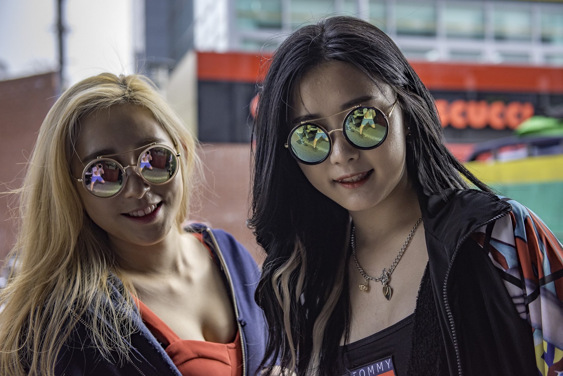 Two young women with sunglasses standing outdoors, reflecting each other in their mirrored sunglasses, with colorful background elements.