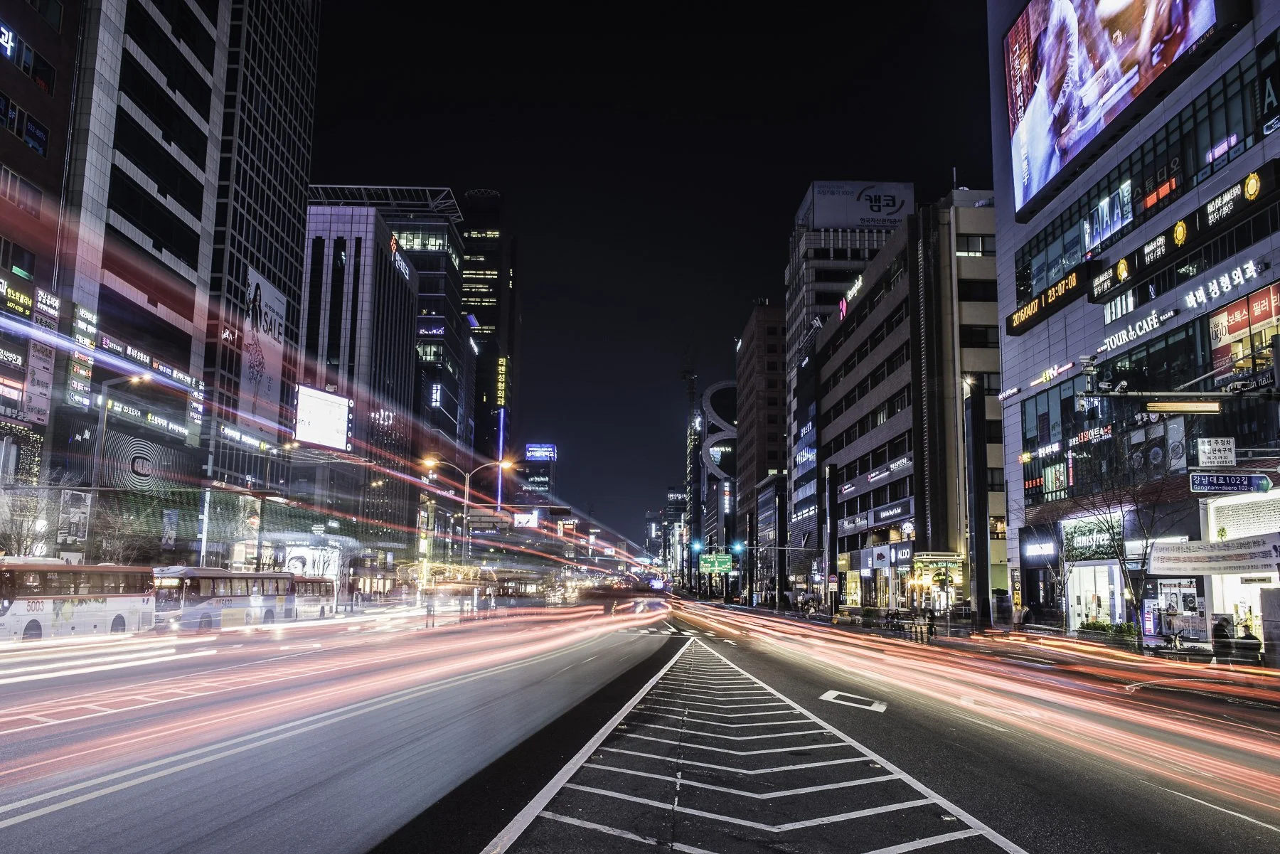 A city street at night with tall buildings, bright neon signs, and light trails from moving vehicles.