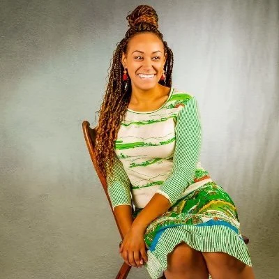 Smiling woman with braids in a colorful green and white dress, sitting on a wooden chair against a gray background.