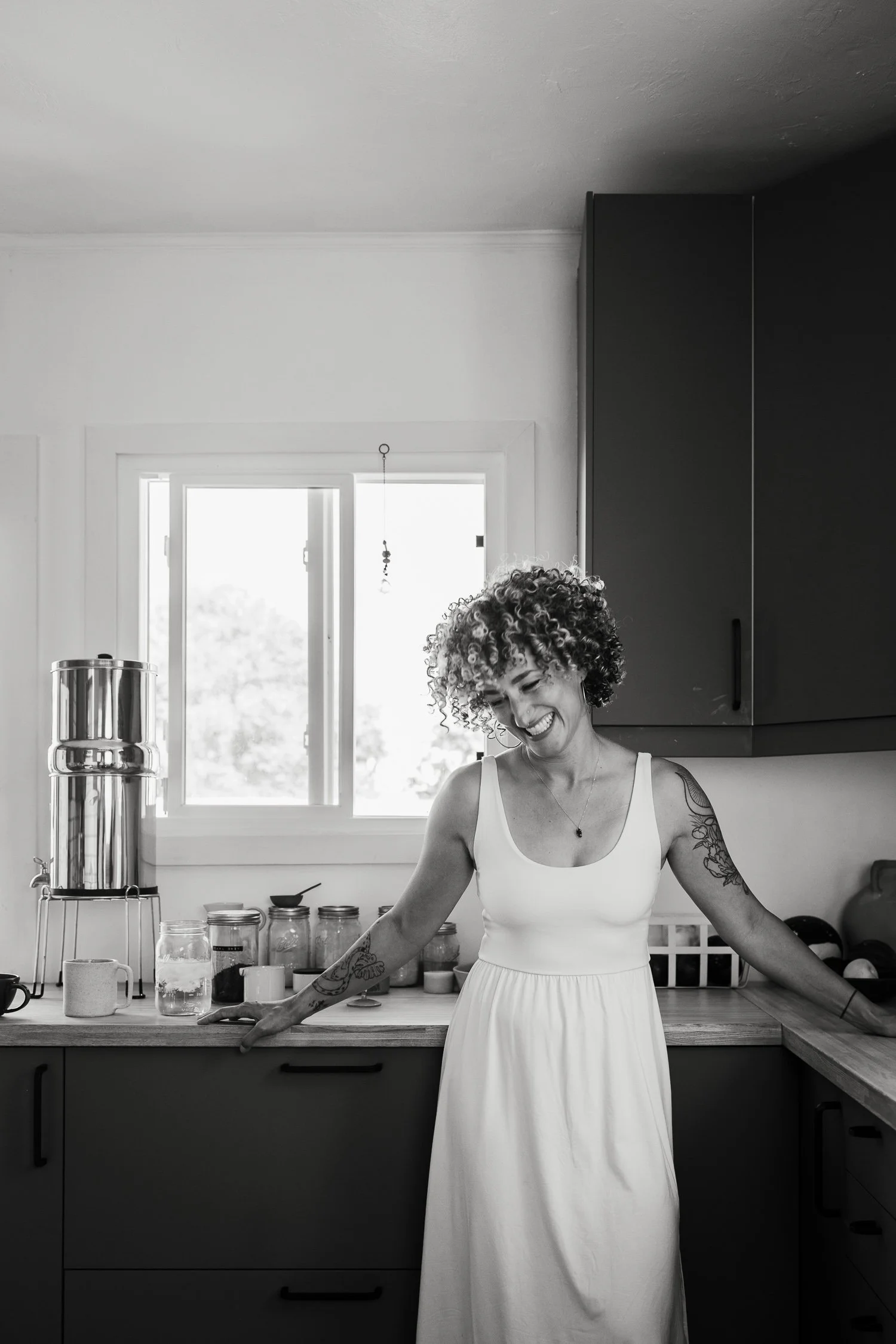 Black and white photo of a woman in a white dress in a kitchen. She is laughing. There is a window behind her.