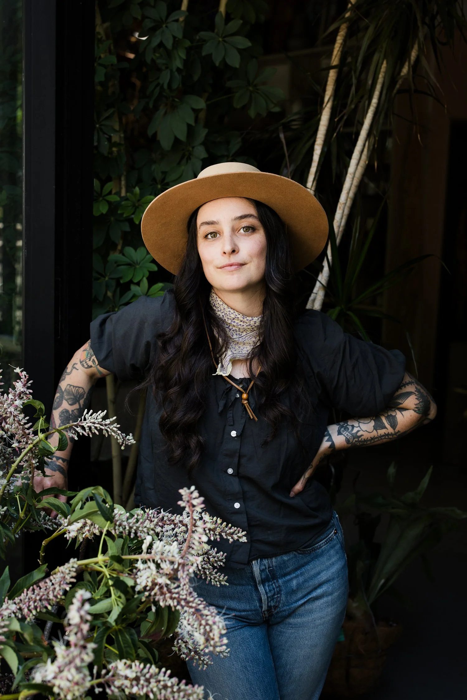 Brand portrait of a florist with tattoos wearing a tan hat and floral scarf standing among lush plants and flowers.