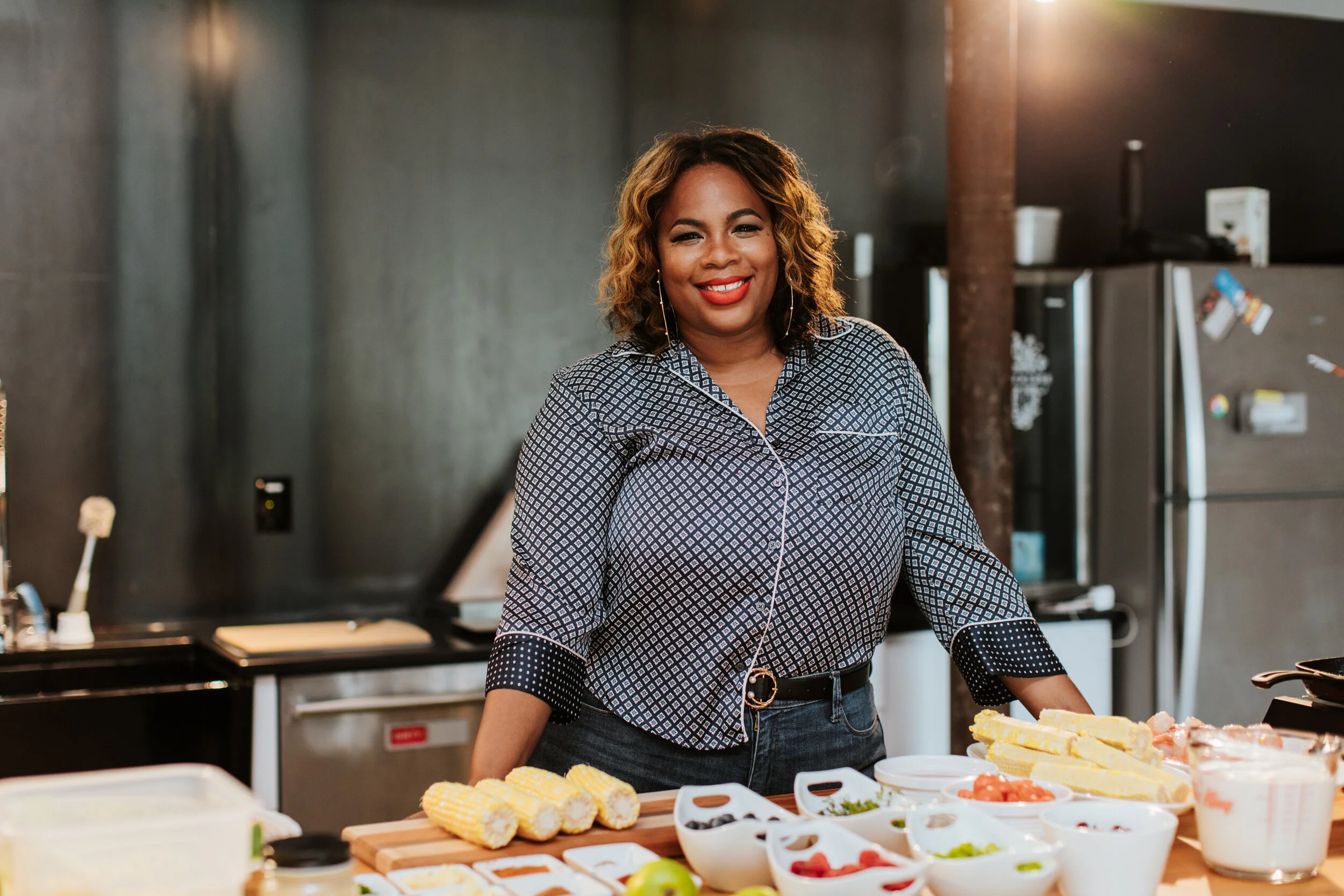 Woman smiling while standing in a kitchen with an assortment of ingredients, including corn, fruits, and vegetables, arranged on a countertop.