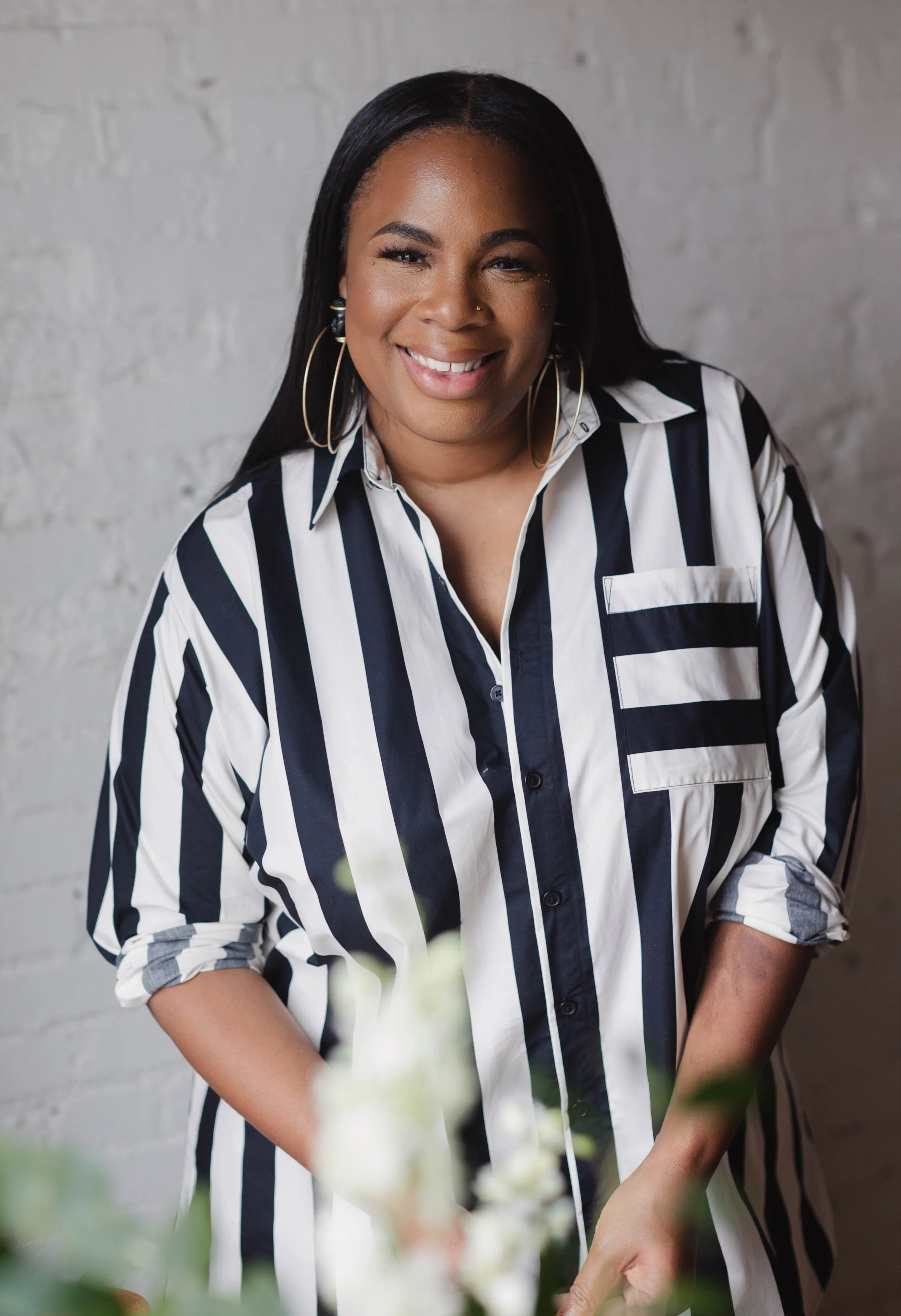 Smiling woman in a striped shirt with large hoop earrings, standing against a white brick wall.