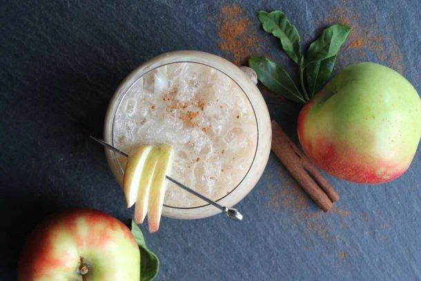 A glass of frothy, apple-flavored beverage garnished with a lemon slice, placed next to two apples, one with green and red coloring, and cinnamon sticks on a dark surface.