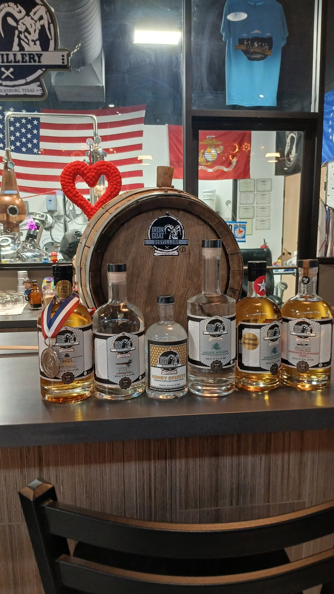 Display of five bottles of alcohol from Iron Goat Distillery on a counter, with a wooden barrel behind them, and flags and T-shirts in the background.