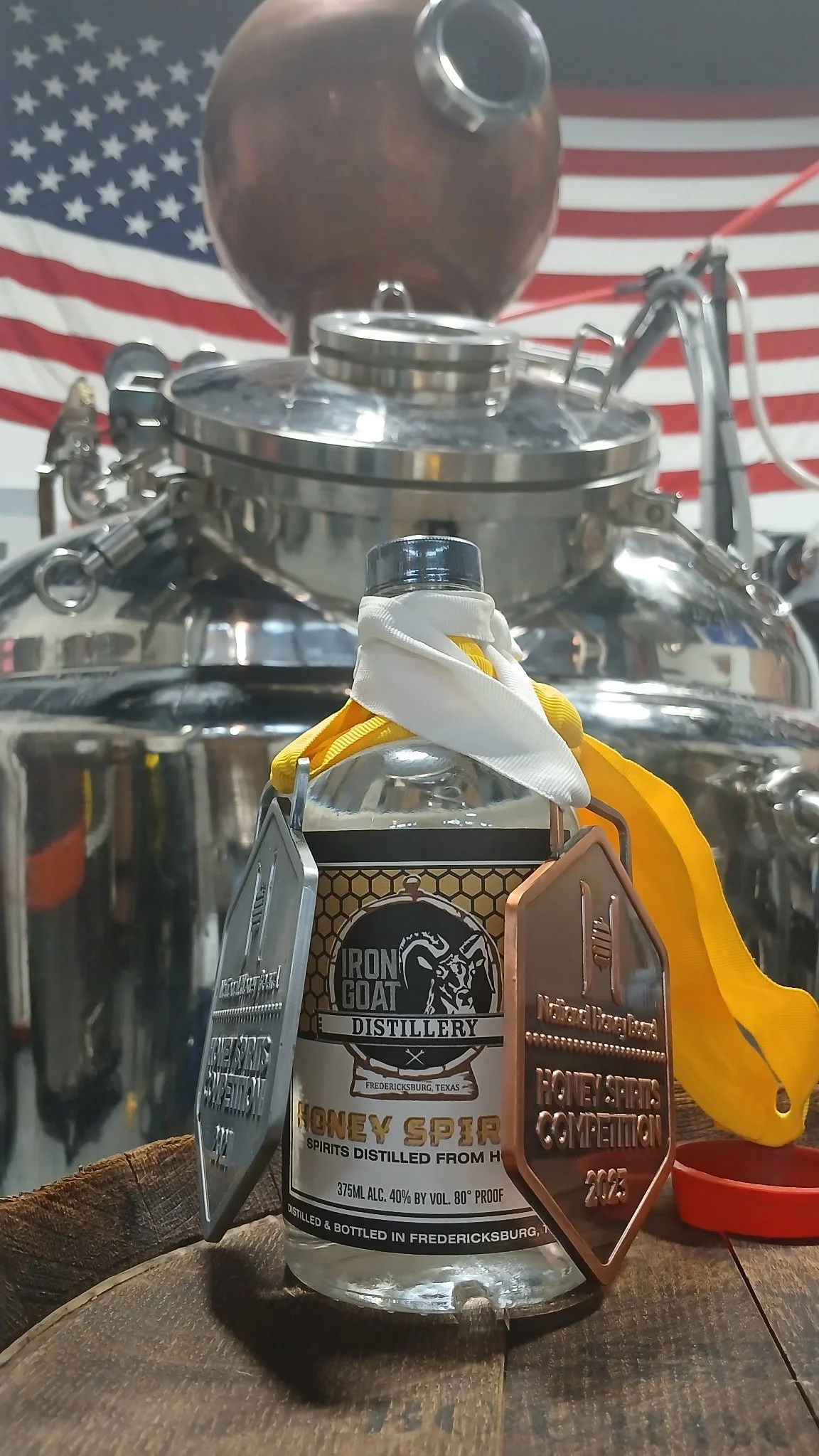 A bottle of honey spirit from Iron Goat Distillery in Fredericksburg, Texas, with a large copper still and American flag in the background.