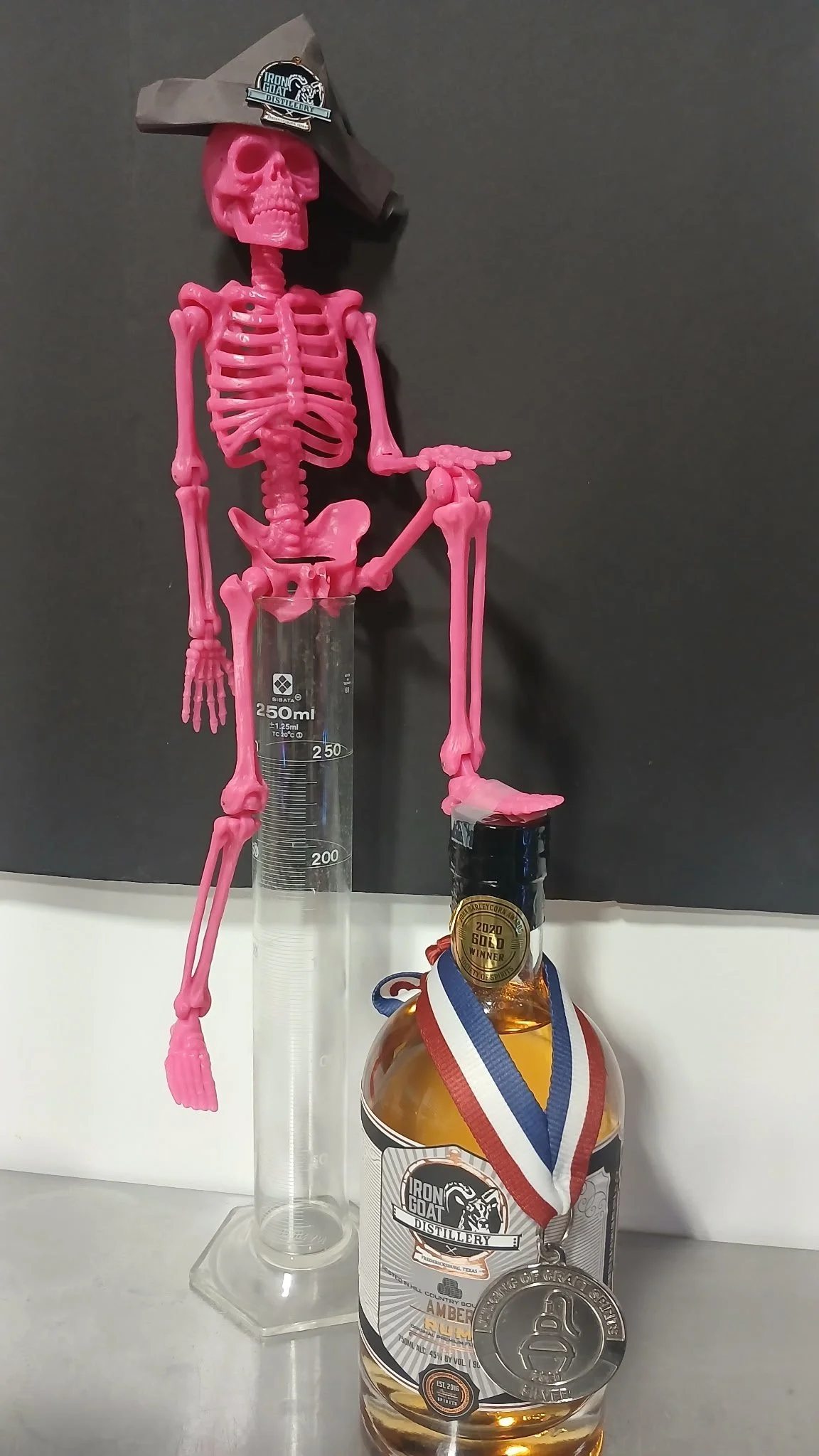 A pink plastic skeleton wearing a Halloween hat with a logo, standing behind a lab beaker with a whiskey bottle and an American flag medal hanging on it. The skeleton appears to be leaning on the bottle, which has an Iron Goat Distillery label and a 