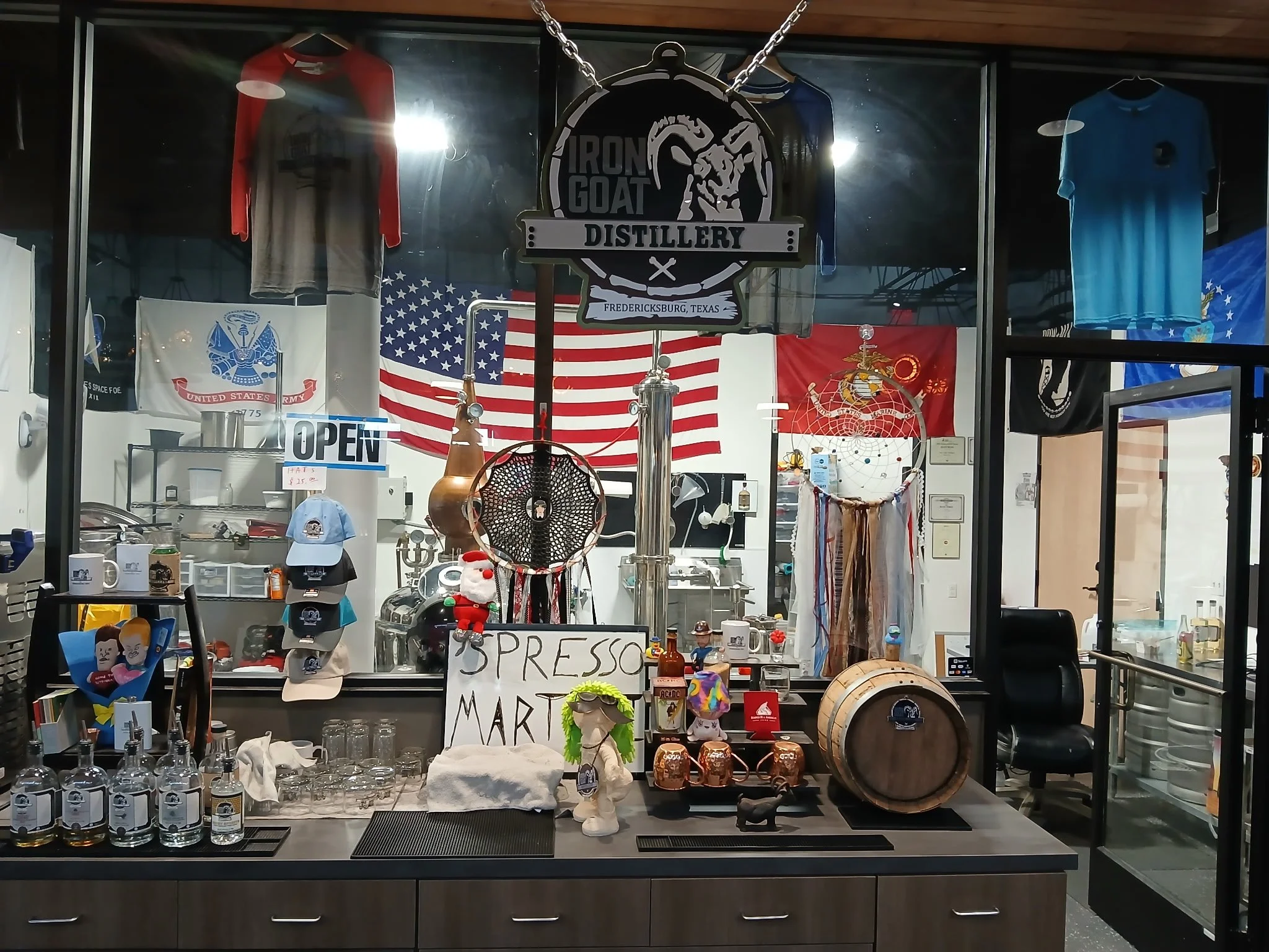 Store window display featuring merchandise including flags, shirts, mugs, hats, glassware, and decorative items, with a sign reading 'Espresso Martini' and a large logo sign for Iron Goat Distillery in Fredericksburg, Texas.