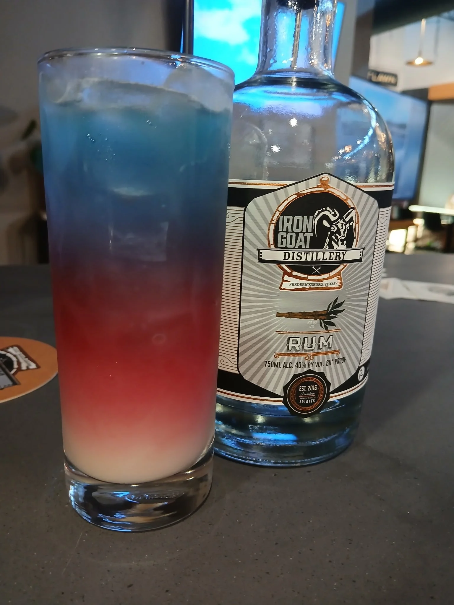 A glass of red, white, and blue layered cocktail sits on a dark surface, with a bottle of Iron Goat Distillery rum in the background. The bottle label indicates it contains rum with 40% alcohol by volume.