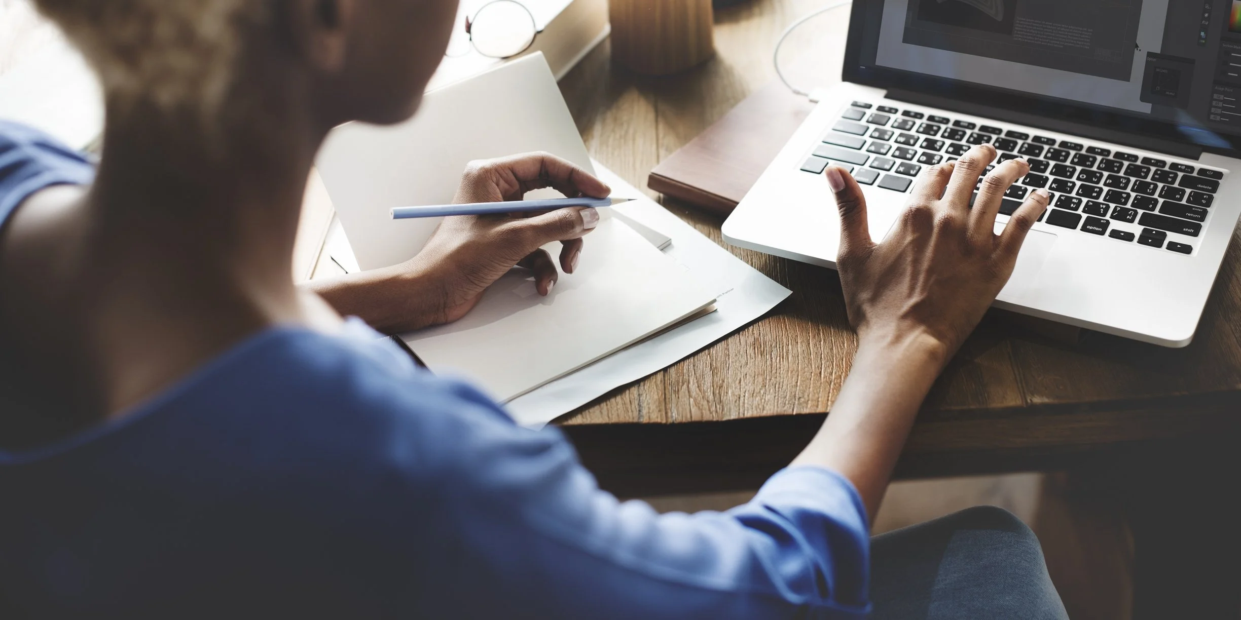 A person using a laptop and taking notes on paper with a pen in a well-lit workspace.
