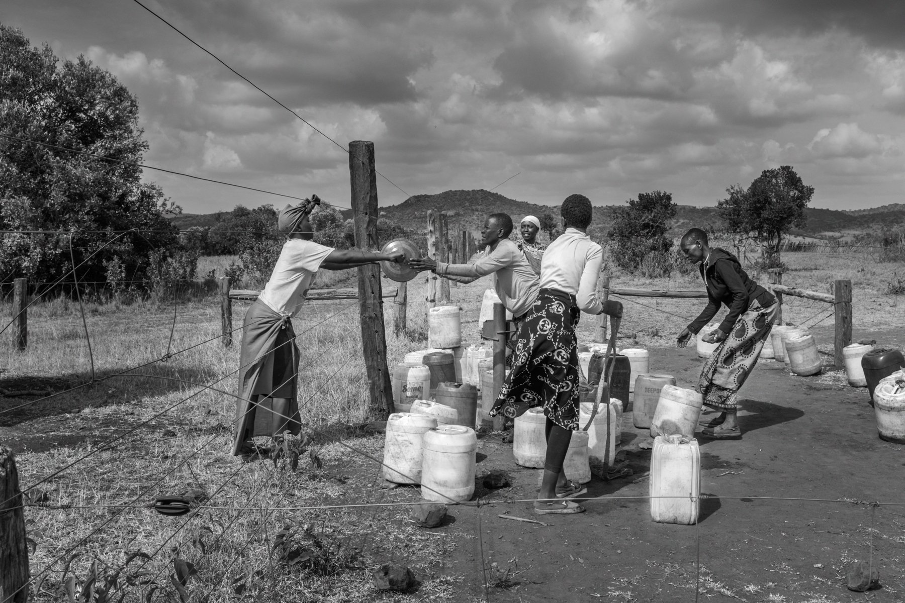  Maasai women gather at the water collection point in their village, close to the fence that separates their community from Borana conservancy. A woman passes a jerrycan through the fence, after which she crawls back form underneath it to the village