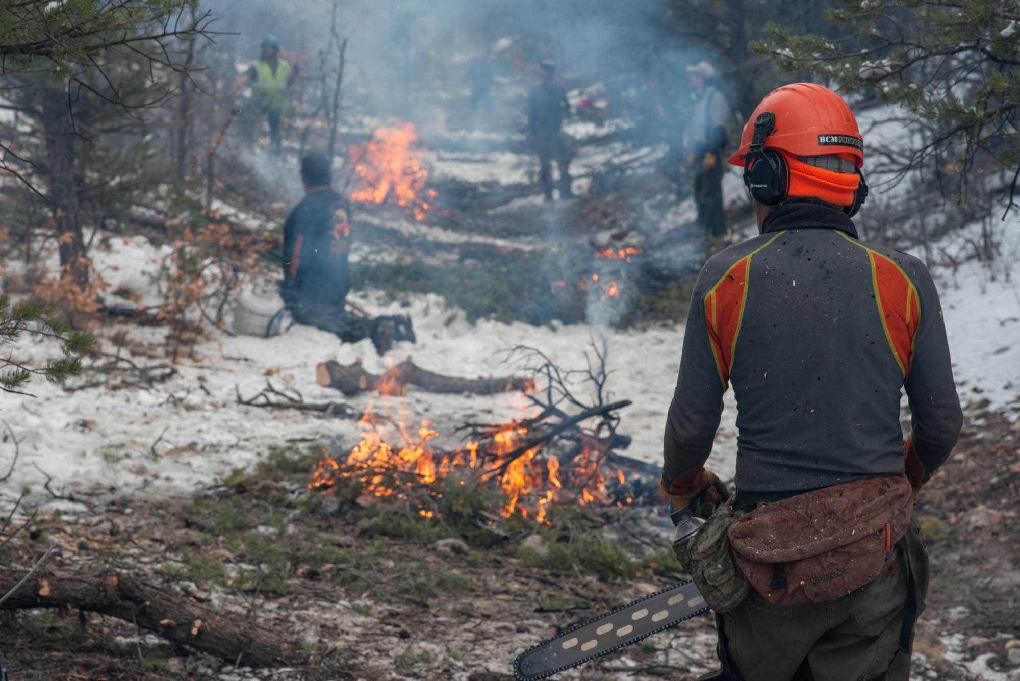  Controlled fires burn in late winter during a thinning mitigation effort to create defensible space. Forester Johnny De’Scoville surveys the scene in the foreground with chainsaw in hand.  