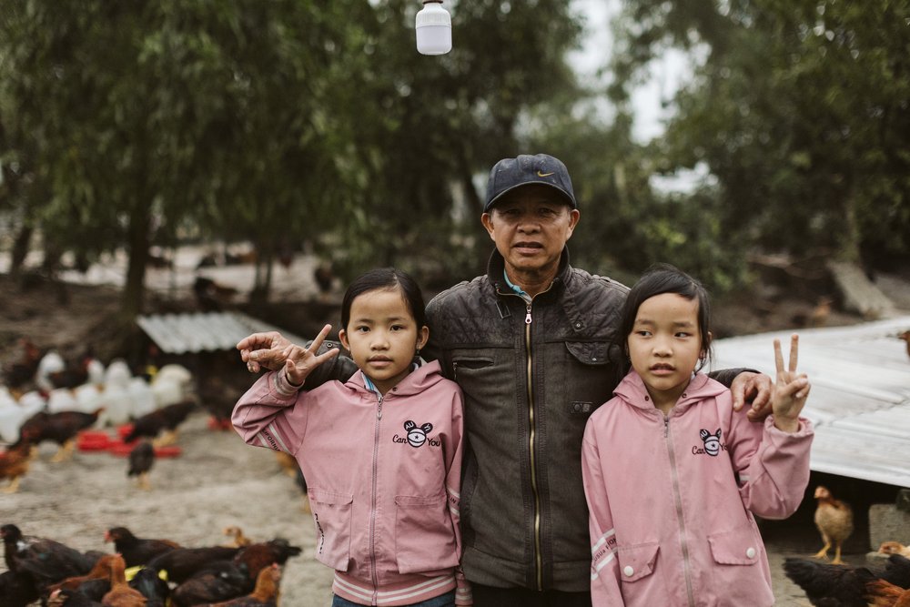 Mr. Tuong is a single father of two girls. Here he stands with his daughters in front of the chicken coop behind their home. Mr. Tuong started with 50 chickens and now raises 3,000 - 5,000 chickens.