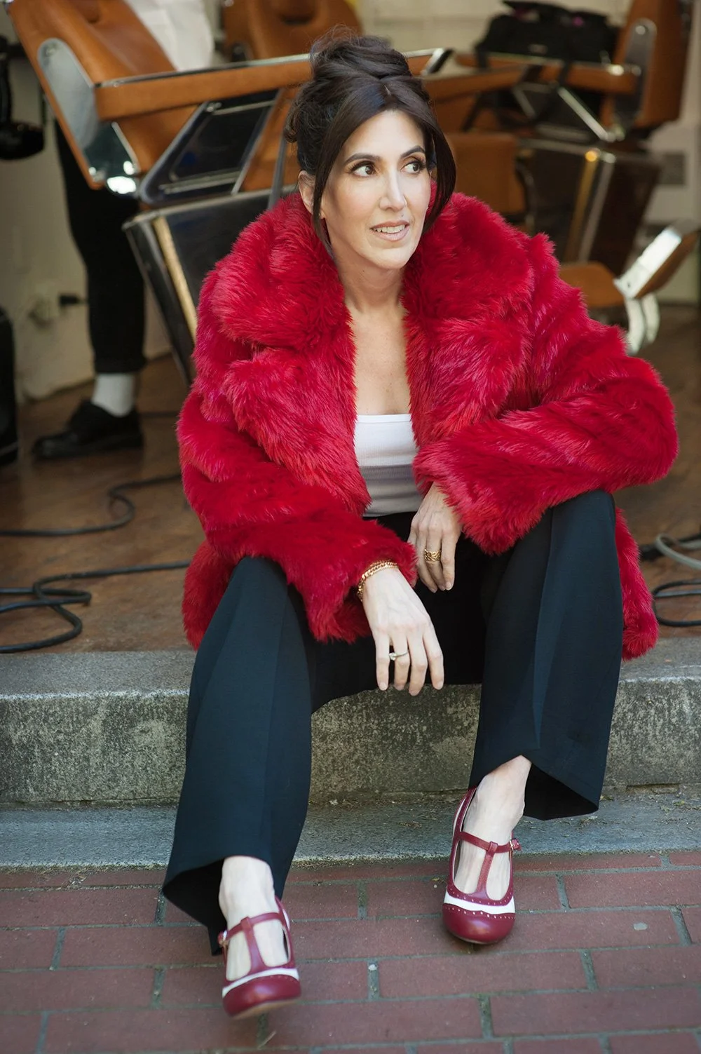 A woman wearing a vibrant red faux fur coat sitting on a concrete step, with black wide-leg pants and red T-strap shoes, sitting outdoors near a building with a stage or performance area behind her.