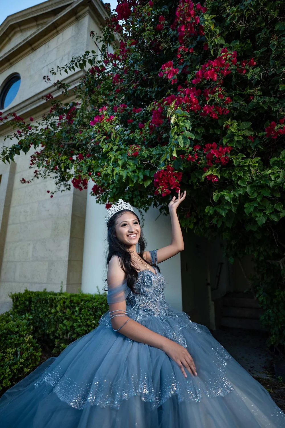 Quinceañera in a blue ball gown and tiara beside red bougainvillea, photographed by Henry M Production in Tampa Bay