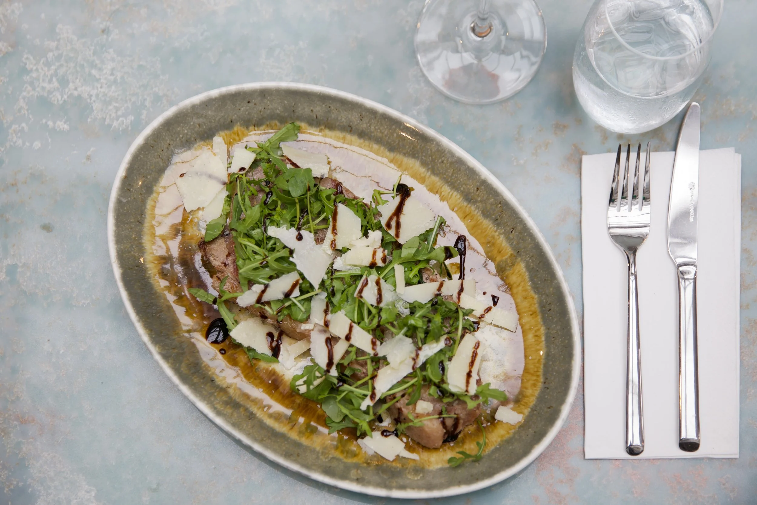 A plate of salad with arugula, shaved cheese, grilled meat, balsamic glaze, olive oil, and other greens on a rustic table. Next to the plate is a fork, knife, a glass of water, and a wine glass.