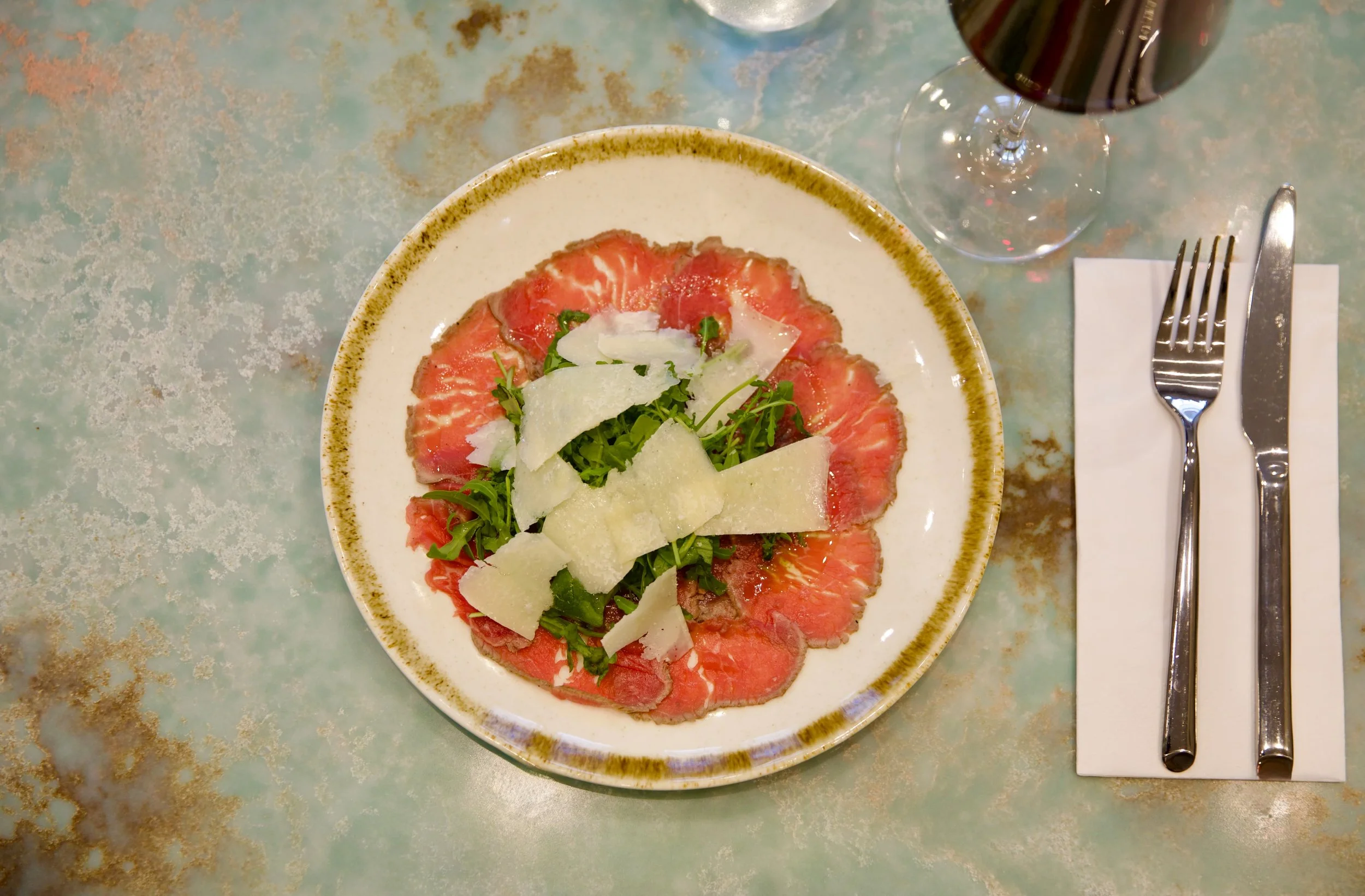 A plate of thinly sliced beef carpaccio garnished with greens and shaved Parmesan cheese, served with a glass of red wine on a table with a fork and knife on a napkin.