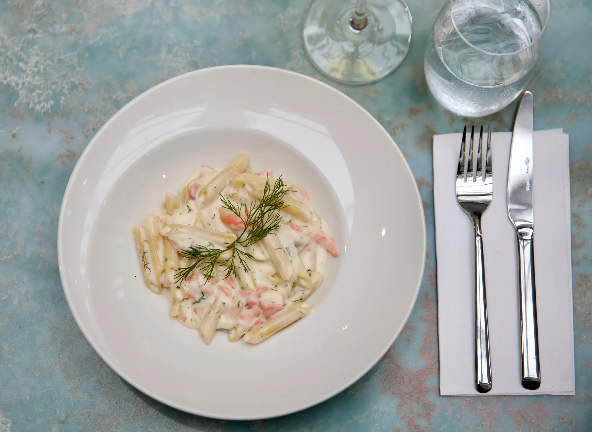 A white plate of creamy penne pasta with pink shrimp and garnished with dill, on a blue distressed table alongside a glass of water, a glass of white wine, a fork, and a knife on a white napkin.