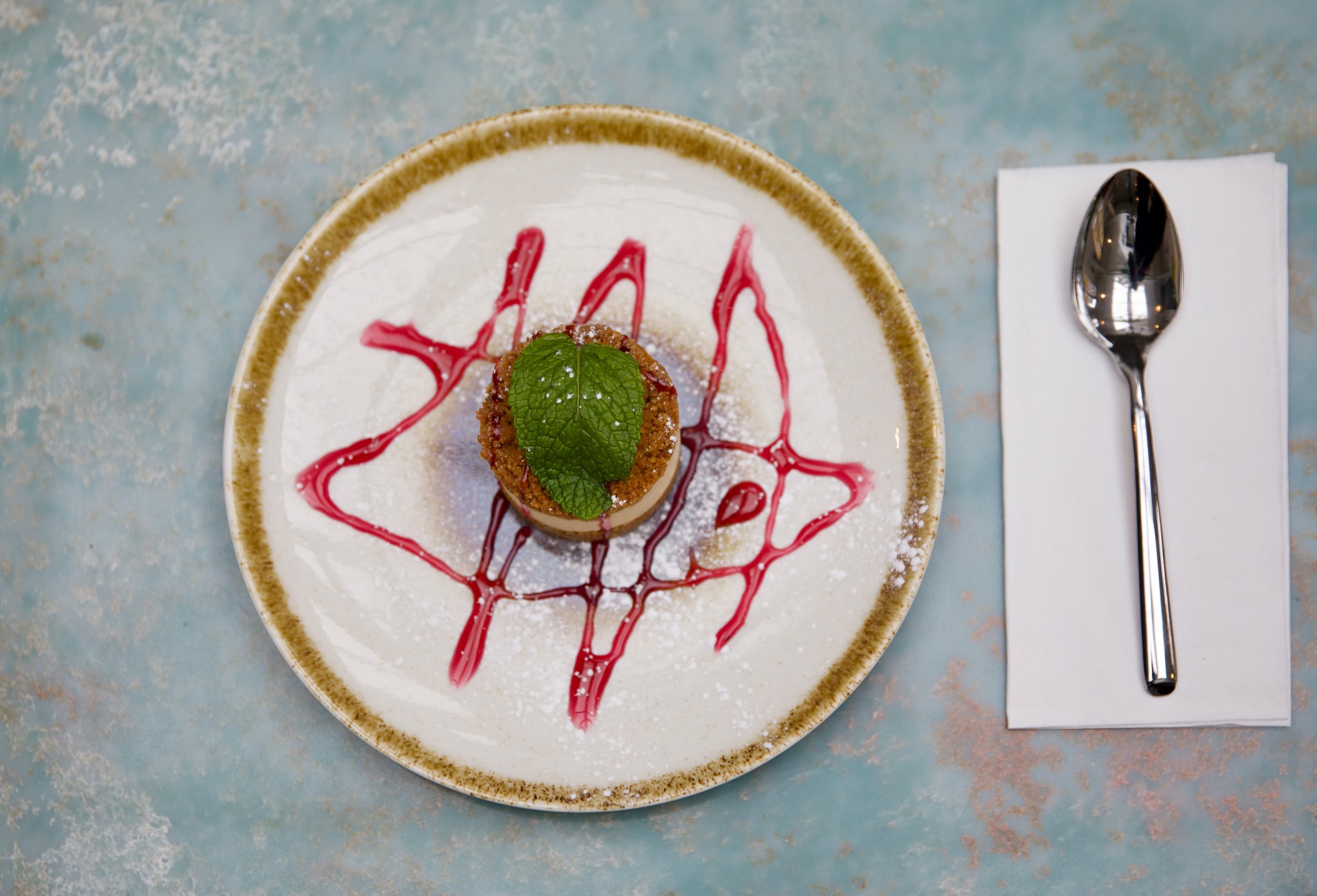  Dessert with a green mint leaf on top, drizzled with red sauce and powdered sugar, served on a rustic plate with a spoon on a napkin.