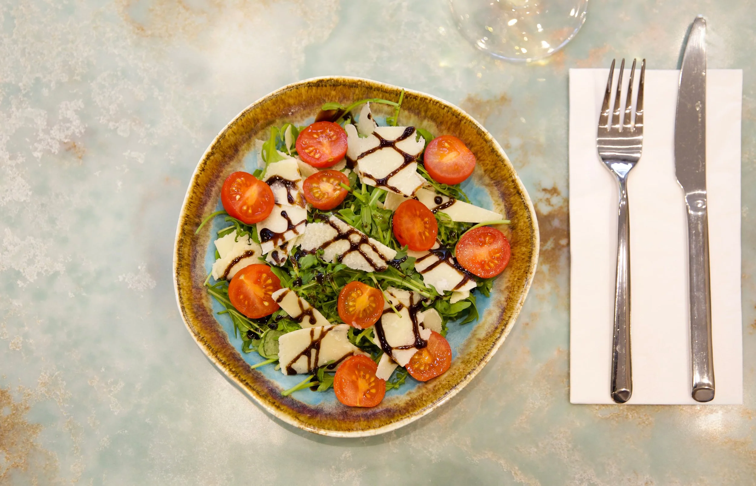 A salad with cherry tomatoes, cheese slices, arugula, and balsamic glaze on a ceramic plate on a table with utensils on a napkin.