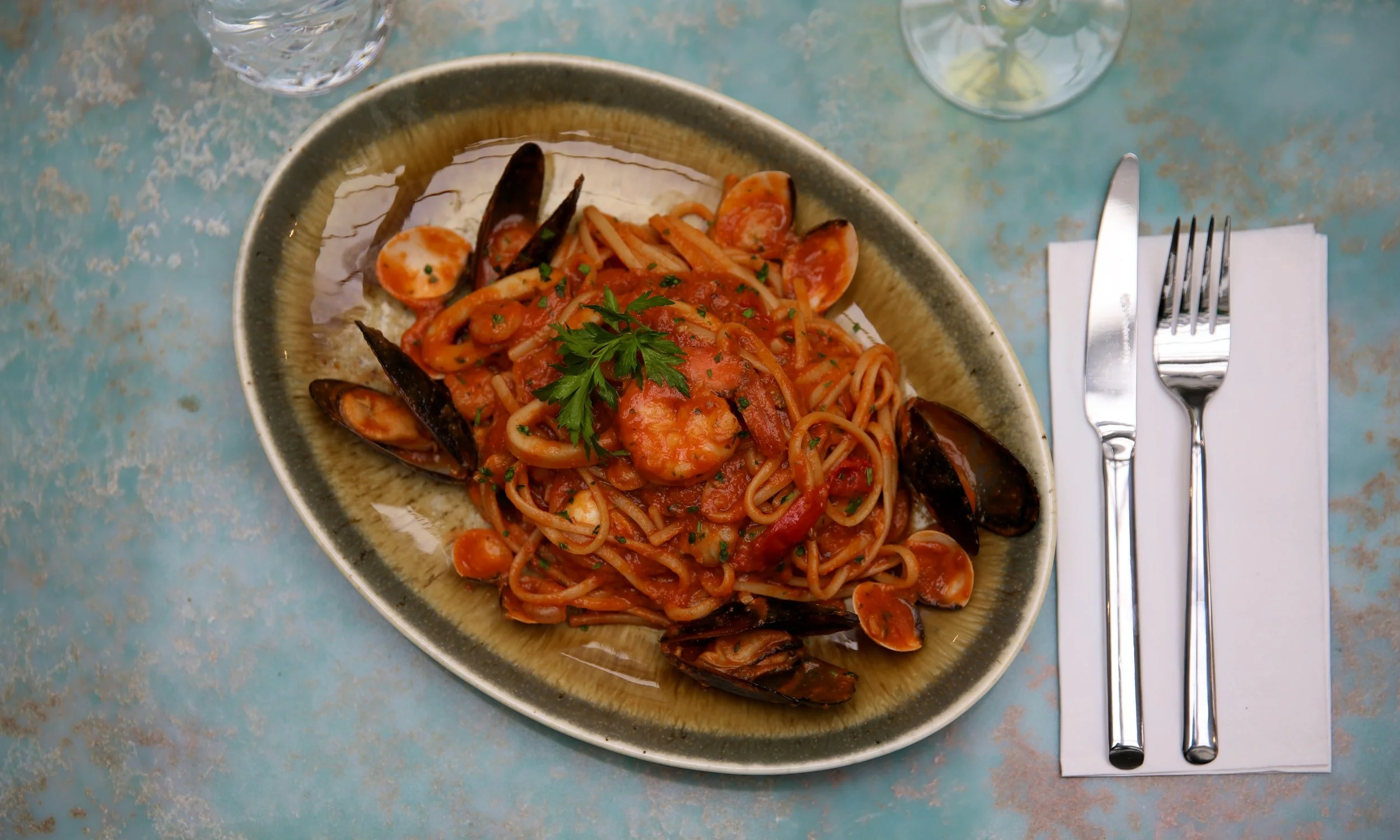 A plate of seafood pasta with squid, mussels, and shrimp garnished with parsley, served on a rustic table with a glass of water and a wine glass.