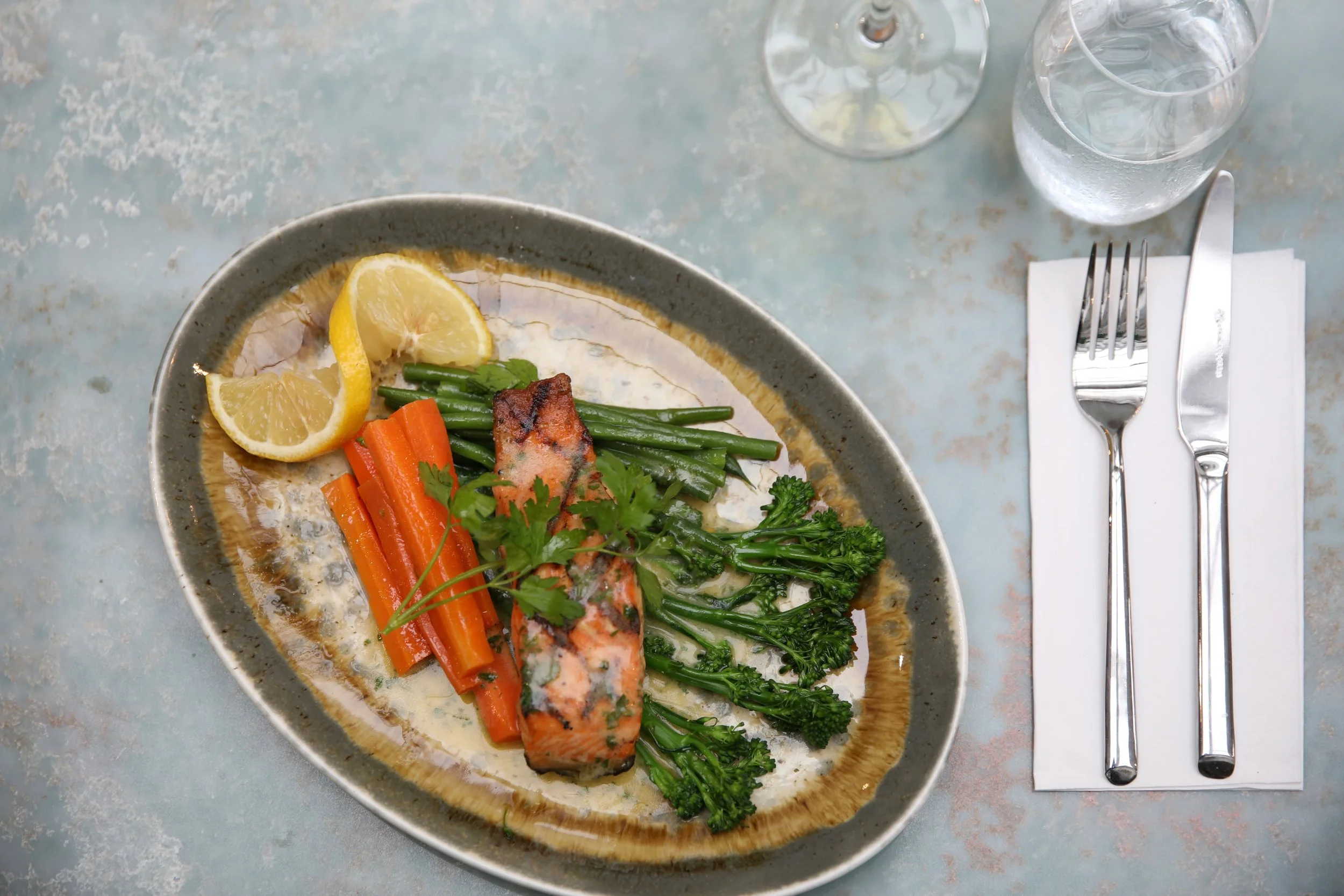 A plate of grilled salmon with lemon wedges, steamed green beans, carrots, broccoli, and cilantro, served with water and set on a table with a fork and knife on a napkin.
