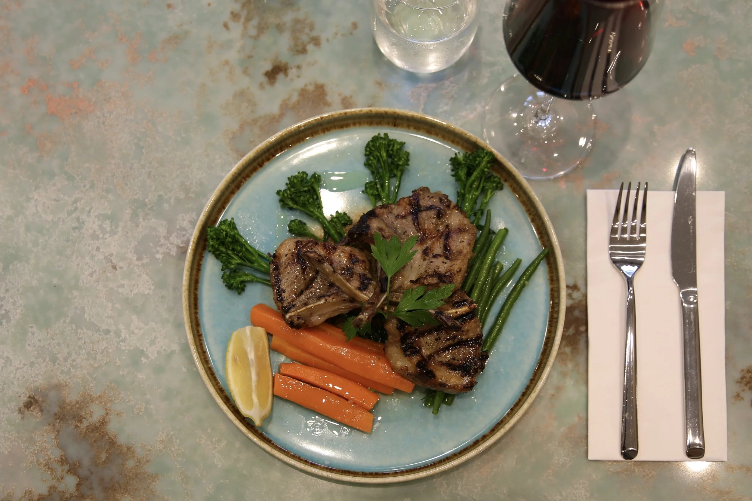 Grilled lamb chops served with green beans, carrots, lemon wedge, parsley garnish, and broccoli on a ceramic plate, with a glass of red wine, a glass of water, and cutlery on a napkin.