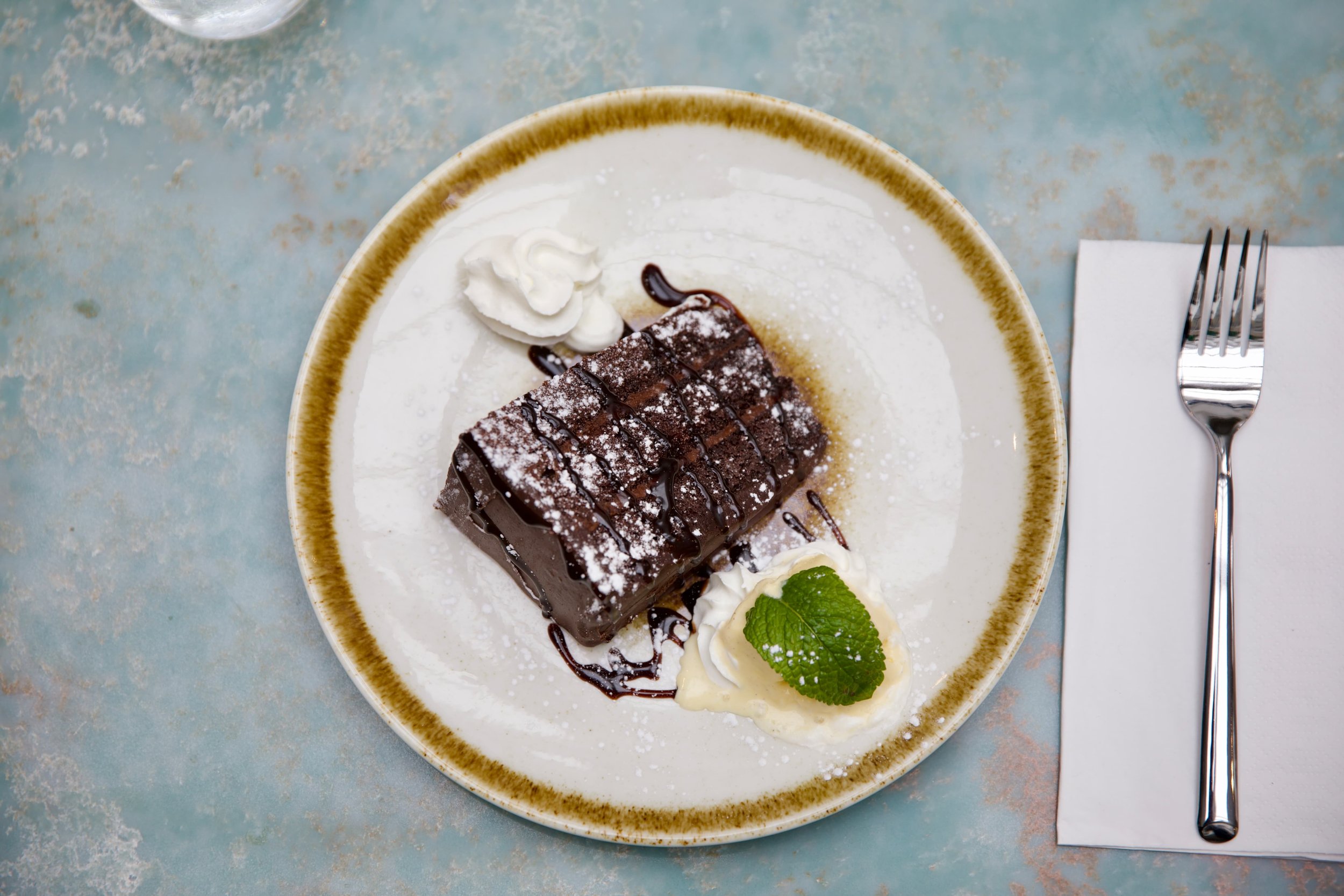 Chocolate lava cake with whipped cream, mint leaf, and chocolate sauce on a white and brown-edged plate.