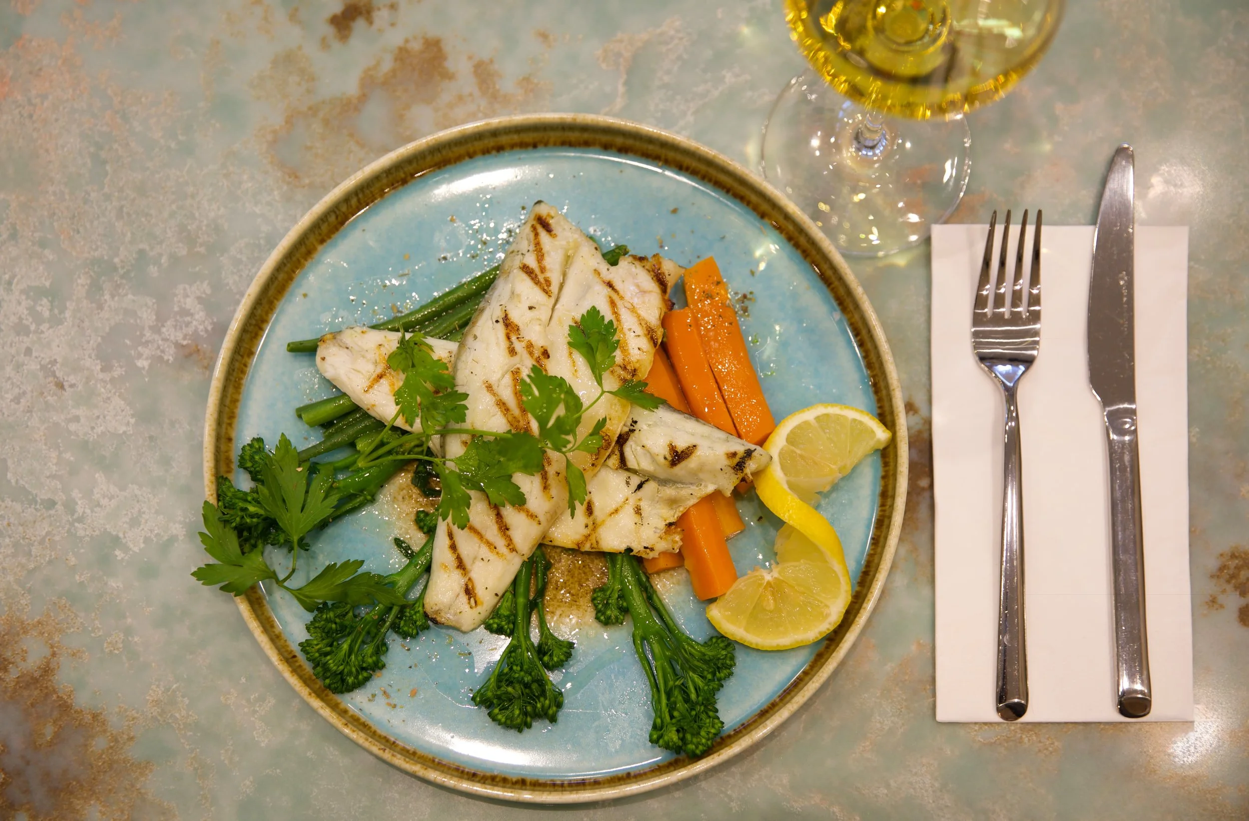 Grilled fish fillet served with green beans, carrots, lemon slices, and fresh herbs on a blue ceramic plate, with a glass of white wine and cutlery on a white napkin.