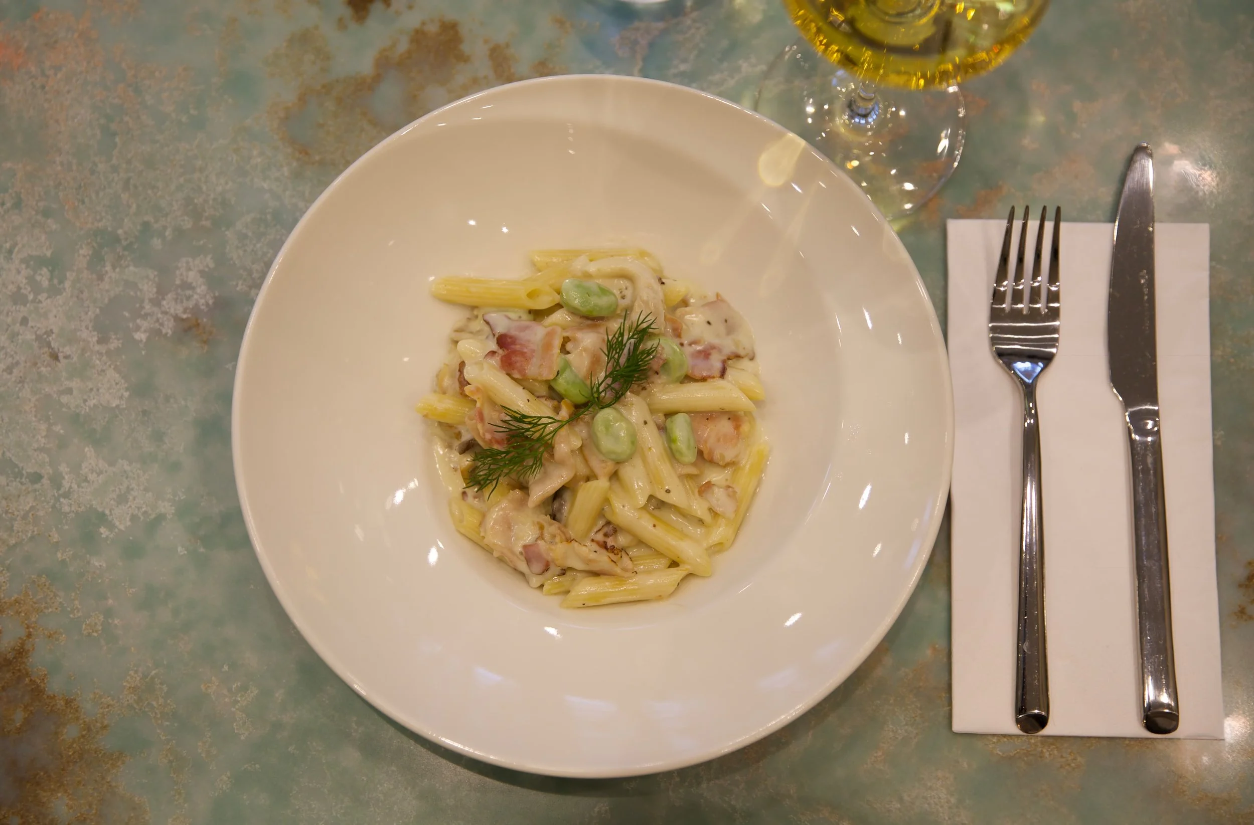 Plate of creamy pasta with green beans and bacon garnished with fresh herbs, with a glass of white wine beside it, placed on a table with a fork and knife on a napkin.