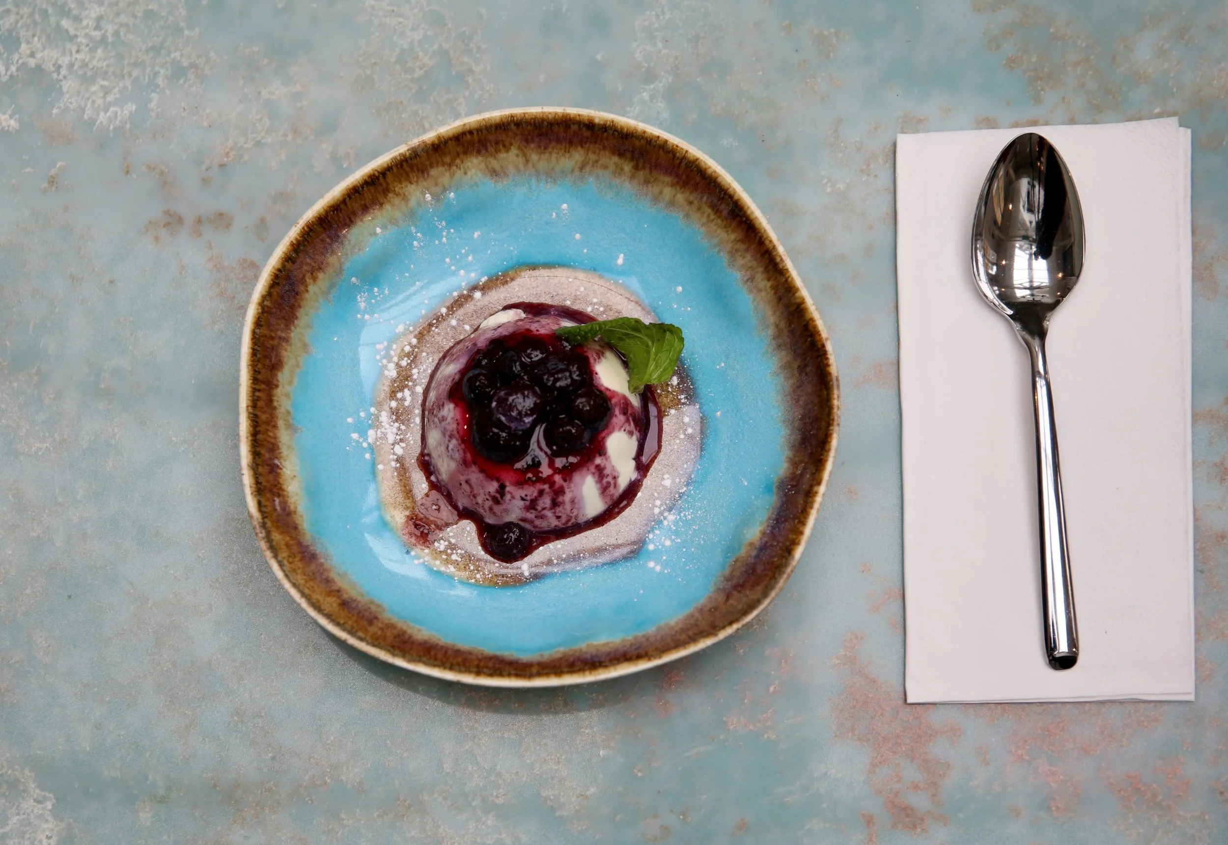 A dessert with blueberries and cream on a blue and brown ceramic plate, garnished with a fresh mint leaf, placed on a textured surface. A metal spoon rests on a white napkin beside the plate.