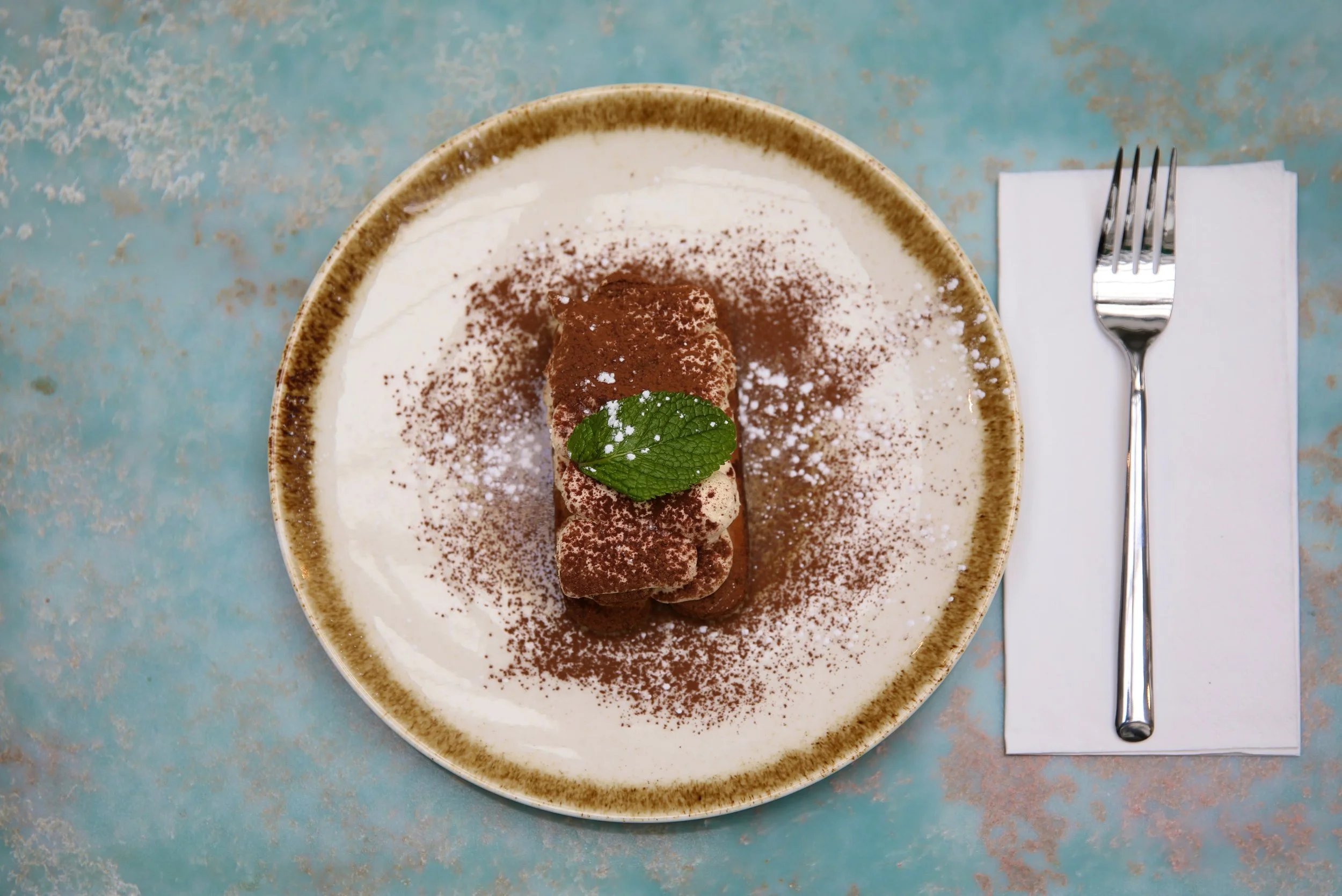 A plated dessert with a rolled cake sprinkled with cocoa powder and a mint leaf, served on a rustic plate with a fork on a white napkin beside it.