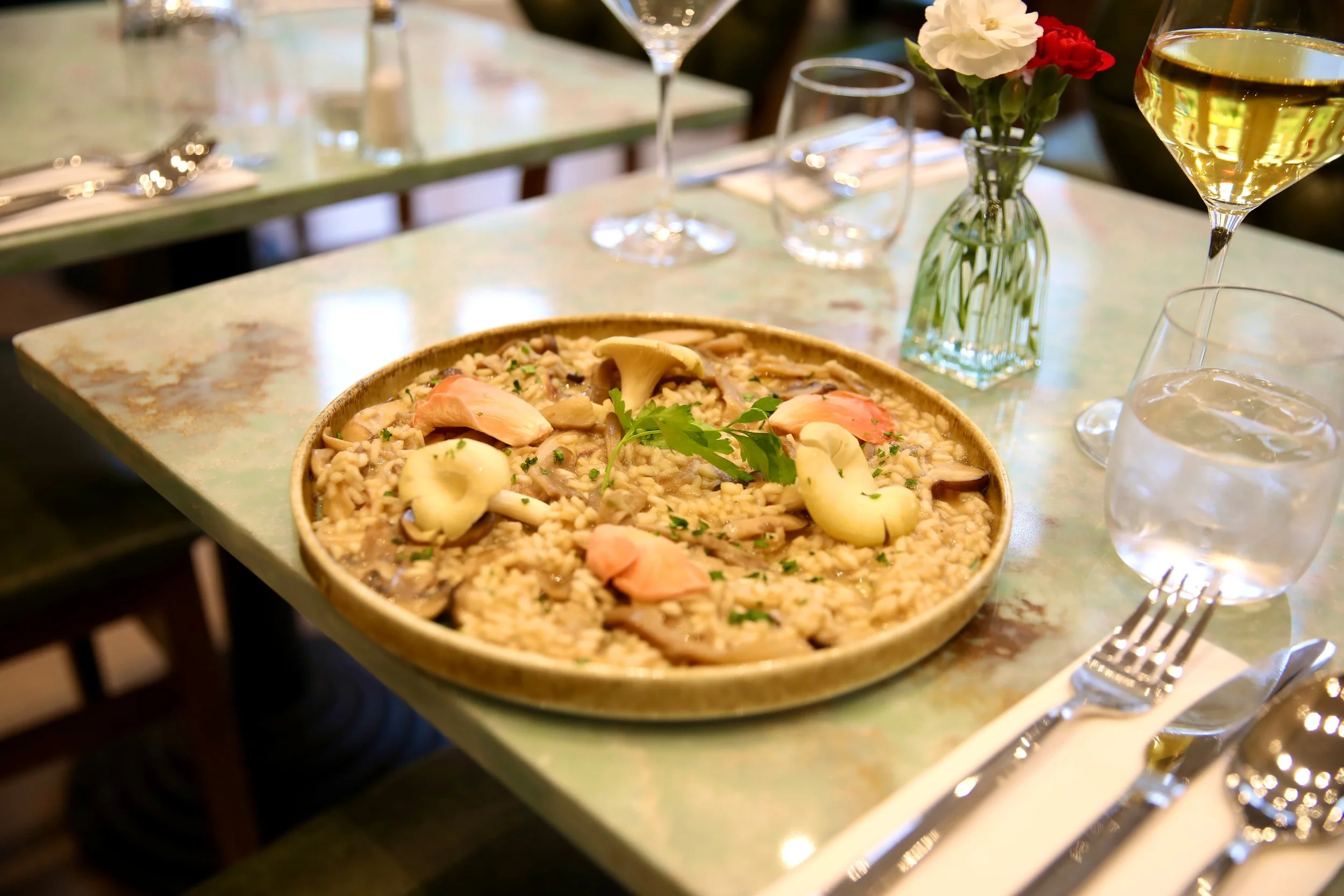 A bowl of mushroom risotto garnished with herbs on a dining table with a glass of white wine, water, a small vase with white and red flowers, and silverware.