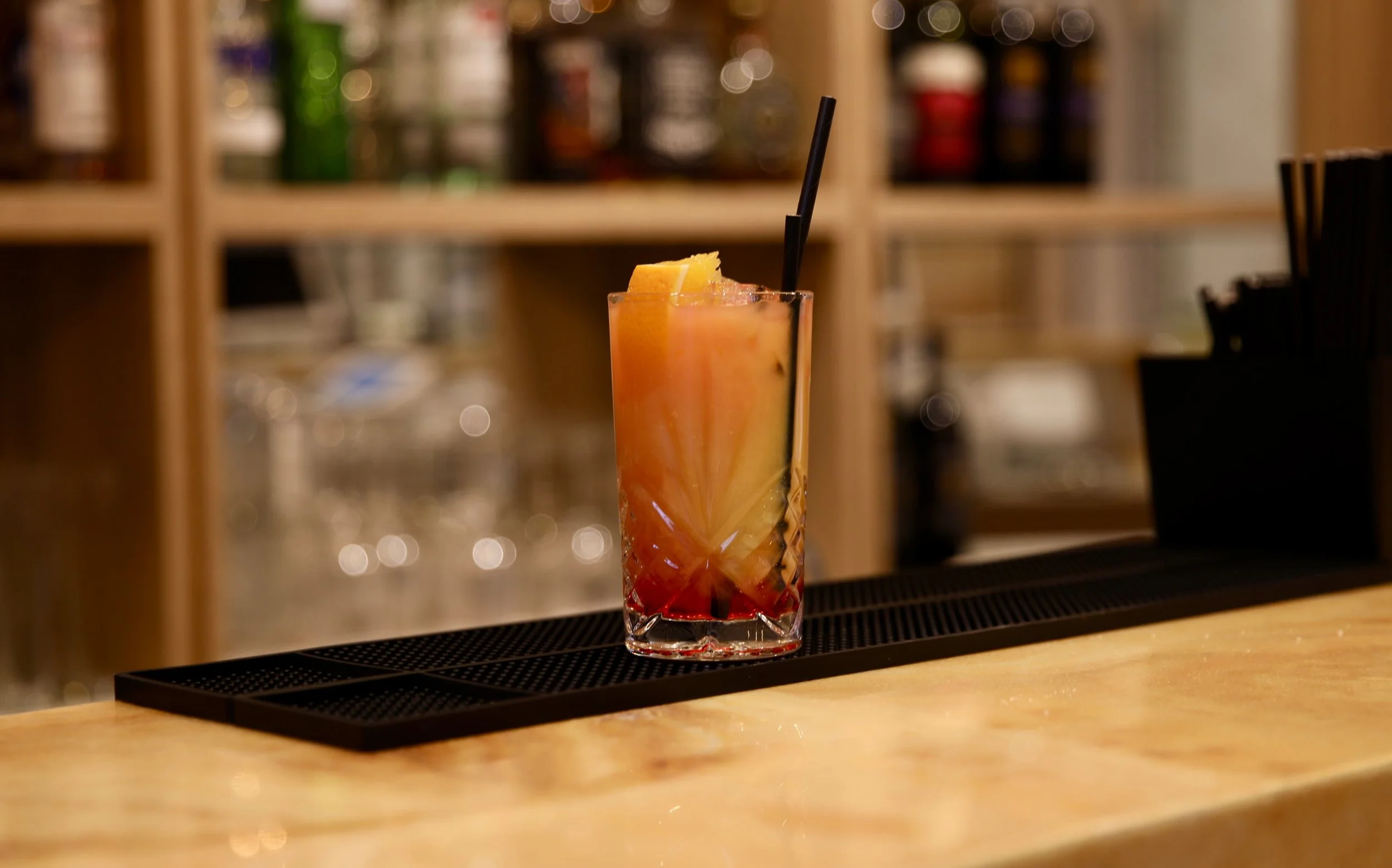 Colorful cocktail with a lemon slice and straw on a bar counter with a blurred bar background.