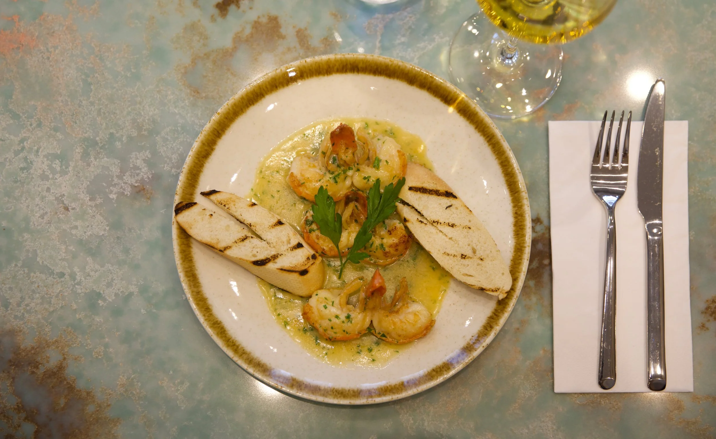 A plate of grilled shrimp with a green sauce, garnished with parsley, accompanied by two pieces of grilled baguette, served on a rustic ceramic plate with a glass of white wine nearby, set on a light green table.