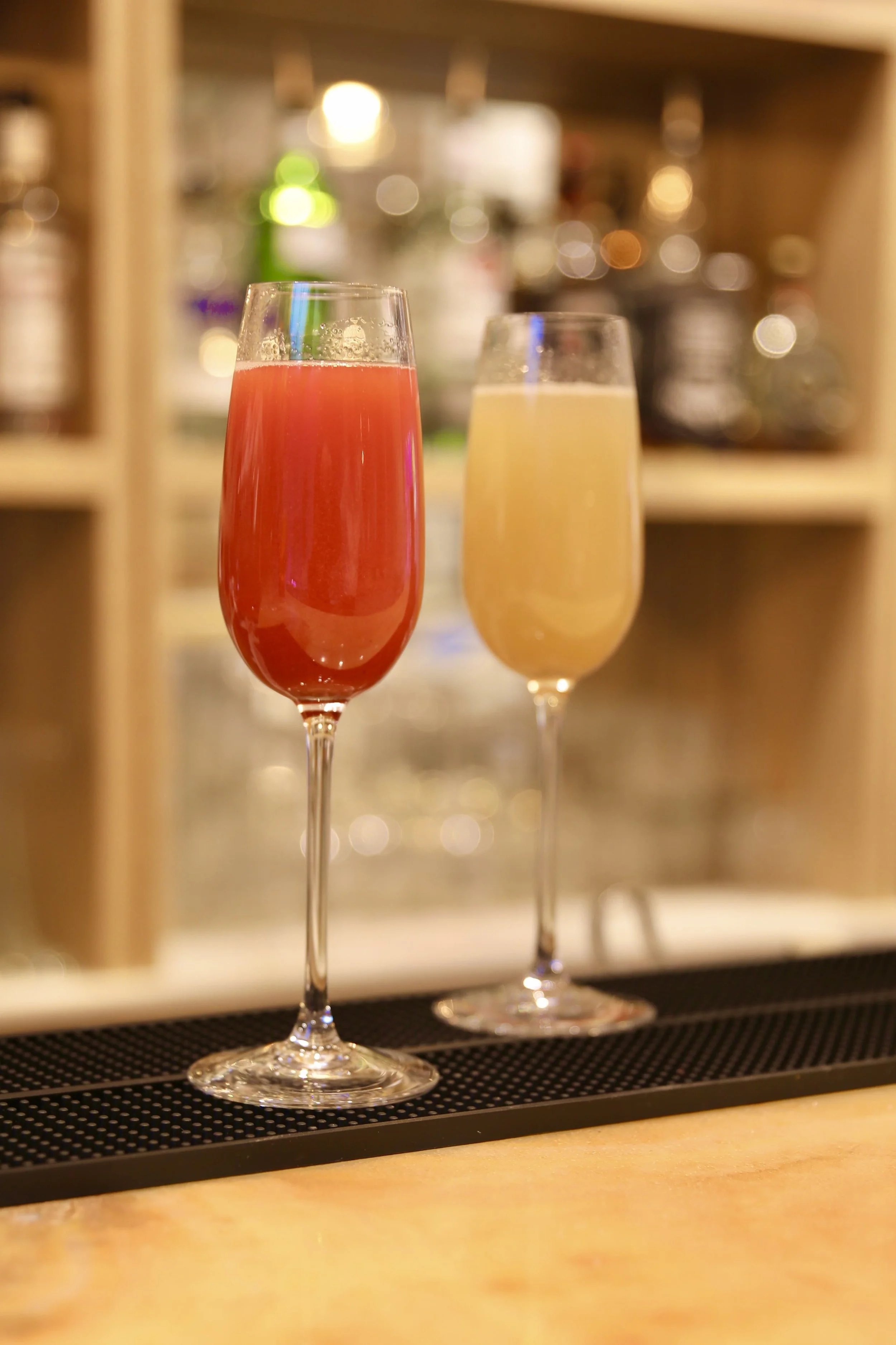 Two champagne flutes, one filled with pink drink and the other with yellow drink, on a bar counter with a blurred background of bottles.