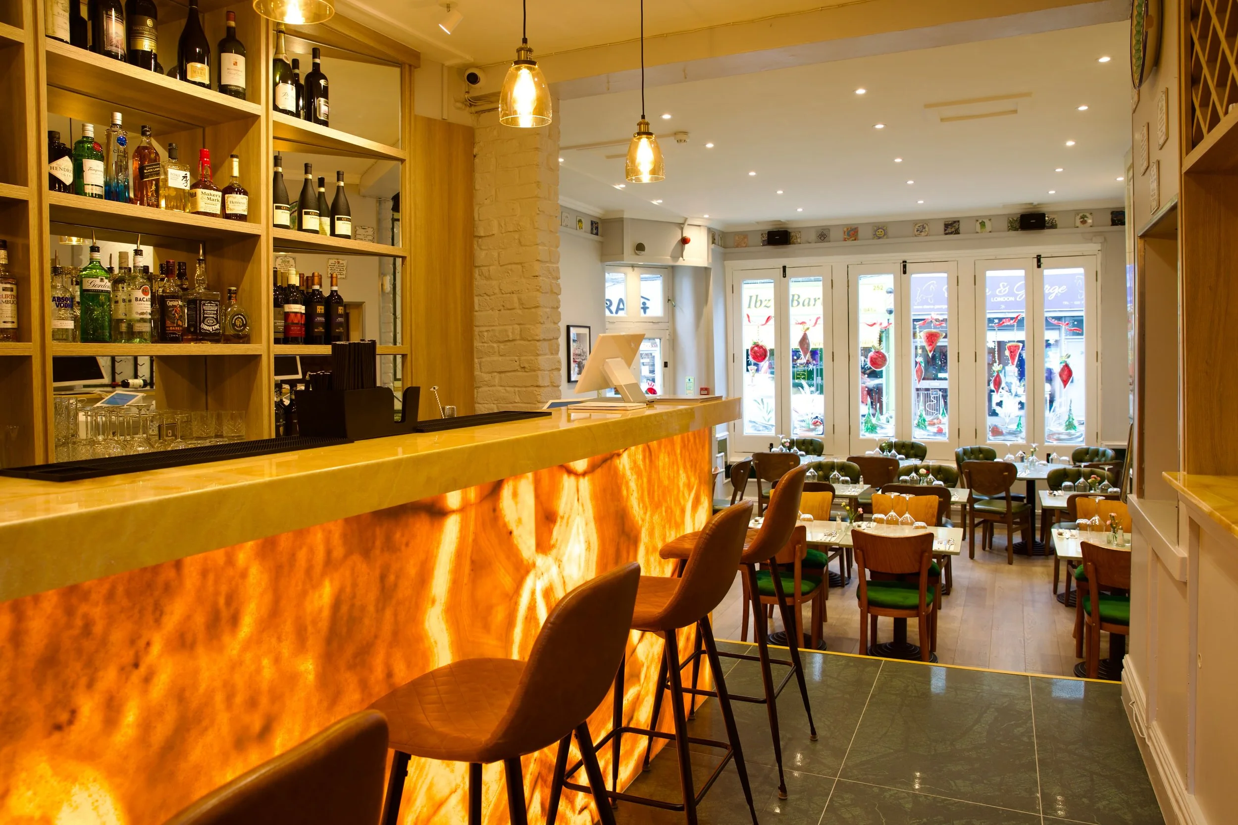 View of a restaurant bar with a glowing amber-colored backlit counter, barstools, and a dining area with tables and chairs near large windows decorated for Christmas.
