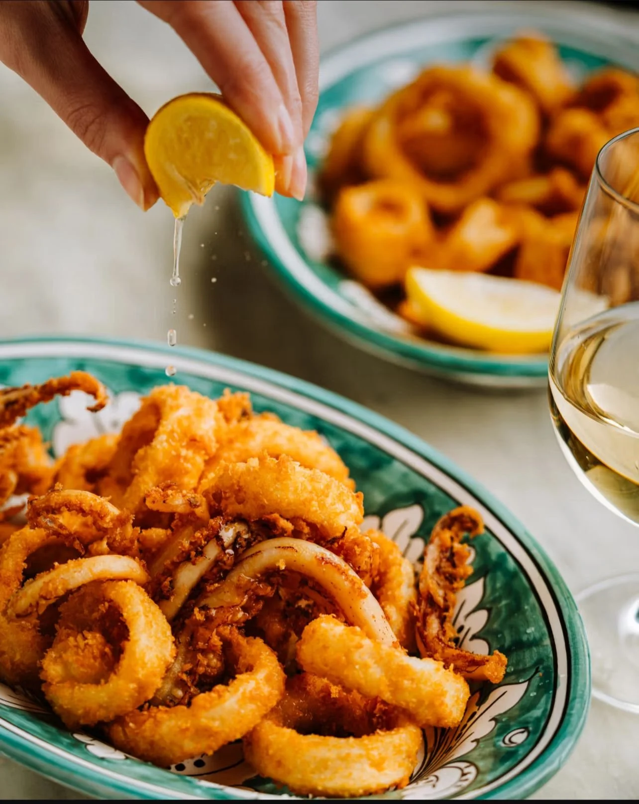 Lemon being squeezed over a plate of fried calamari with a glass of white wine nearby.