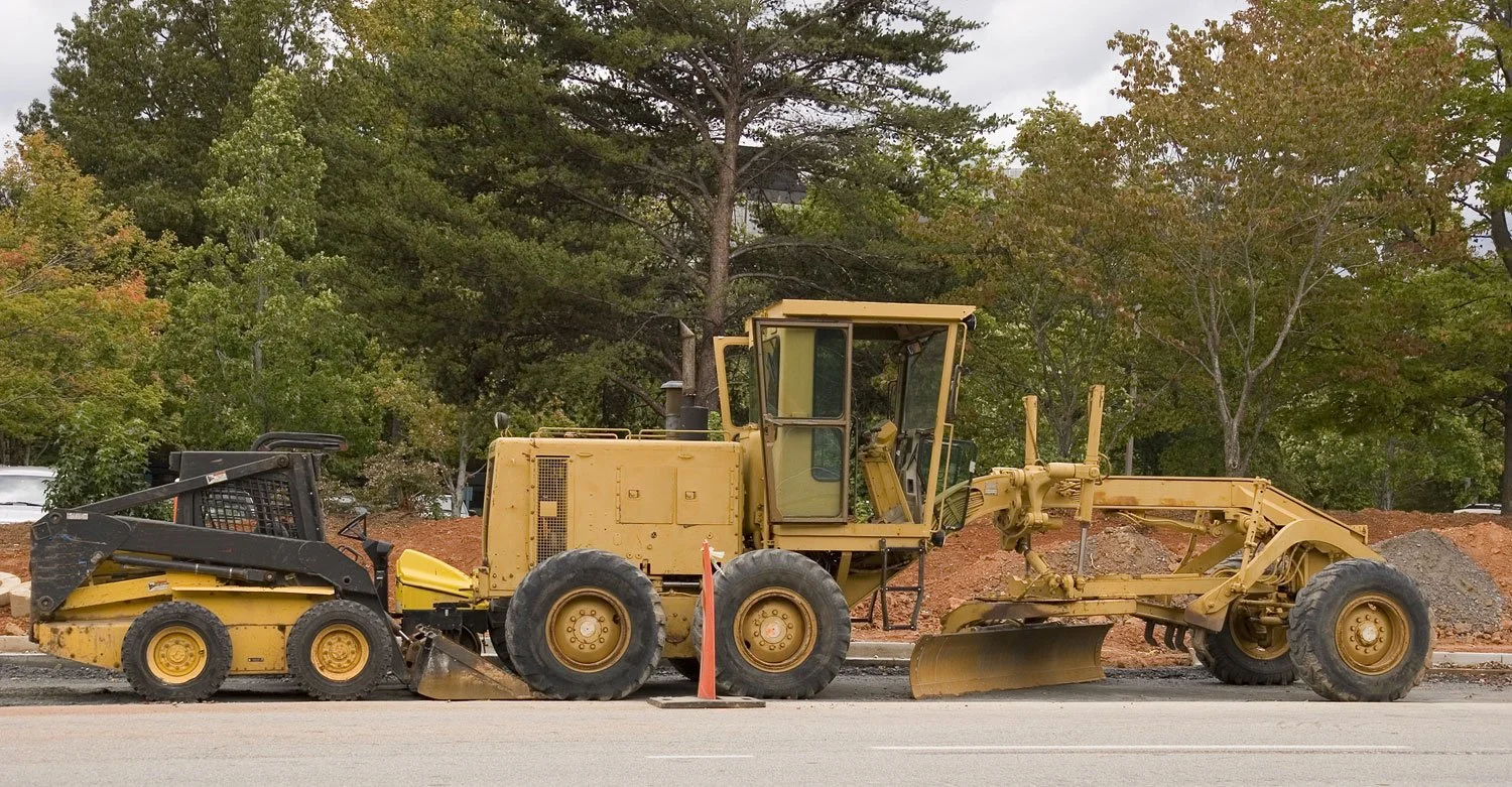 Yellow road grader and skid steer parked at a road construction site with piles of dirt and trees in the background.jpg