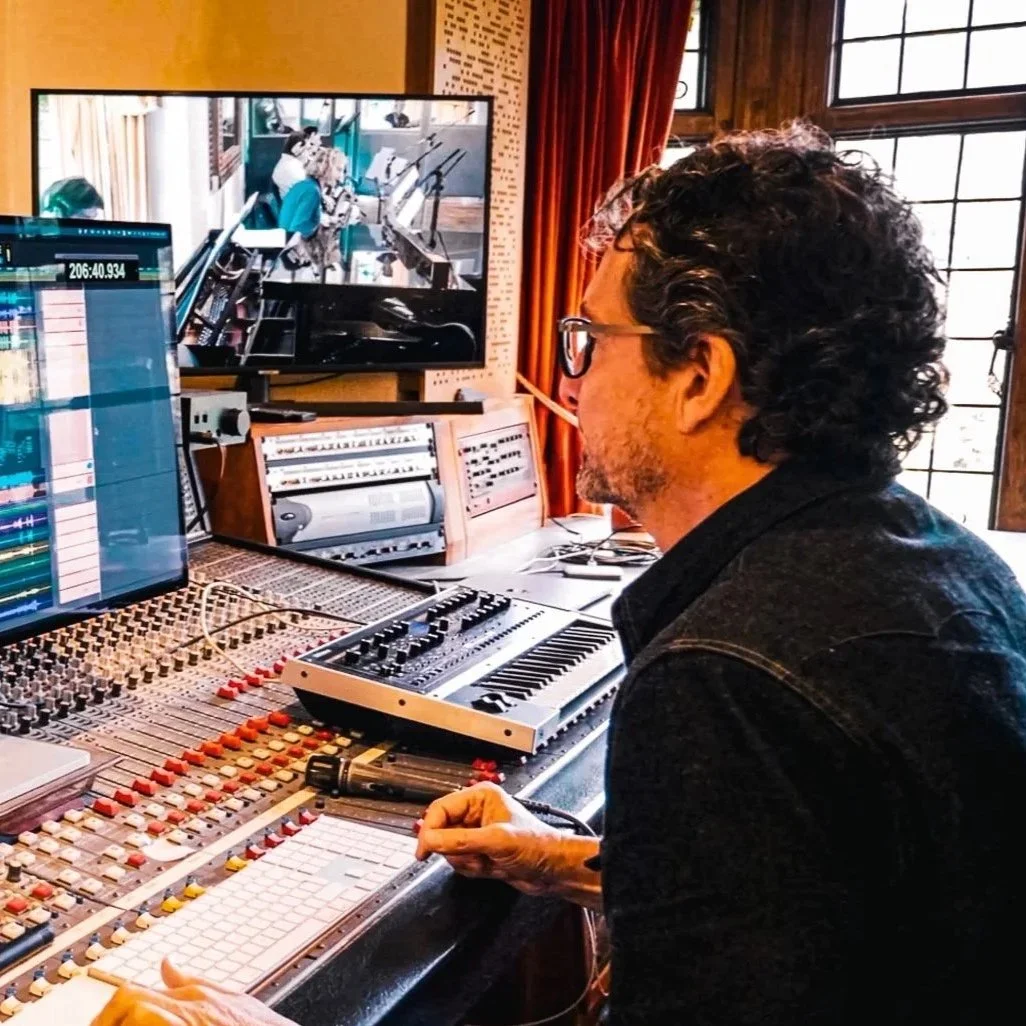 A man with curly hair and glasses working in a recording studio, sitting at a mixing console with multiple colorful knobs and sliders, with monitors displaying audio editing software, and a large window with red curtains nearby.