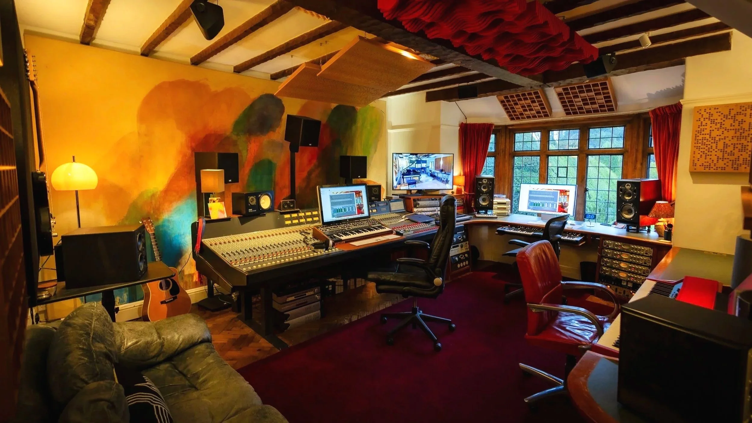 Music recording studio with mixing console, computers, monitors, speakers, musical instruments, and acoustic treatment in a cozy, well-lit room with large windows and colorful wall art.