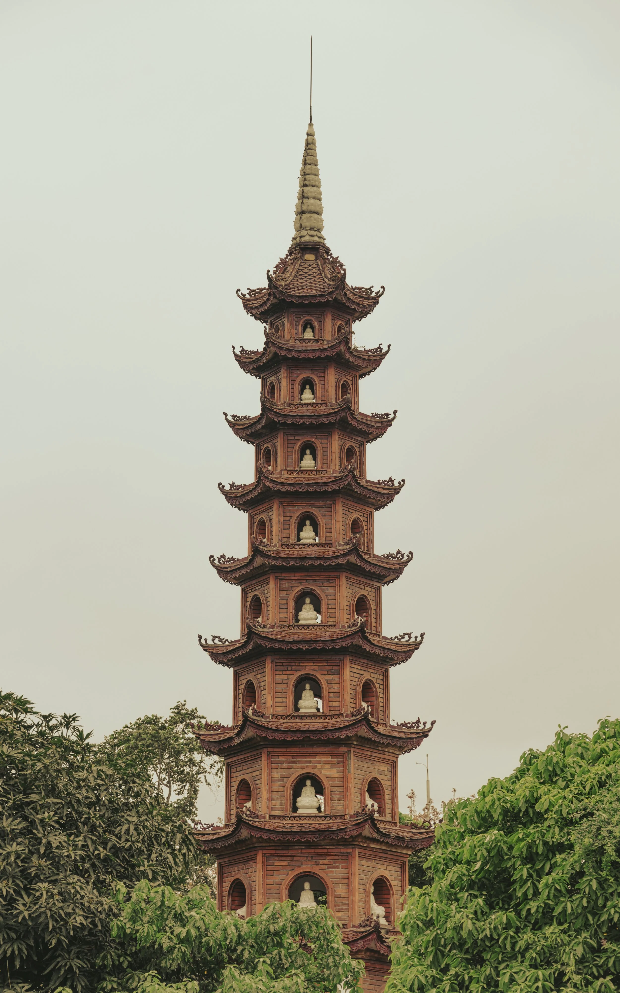 A tall, traditional Asian pagoda with multiple tiers, each featuring small Buddha statues, surrounded by green foliage.