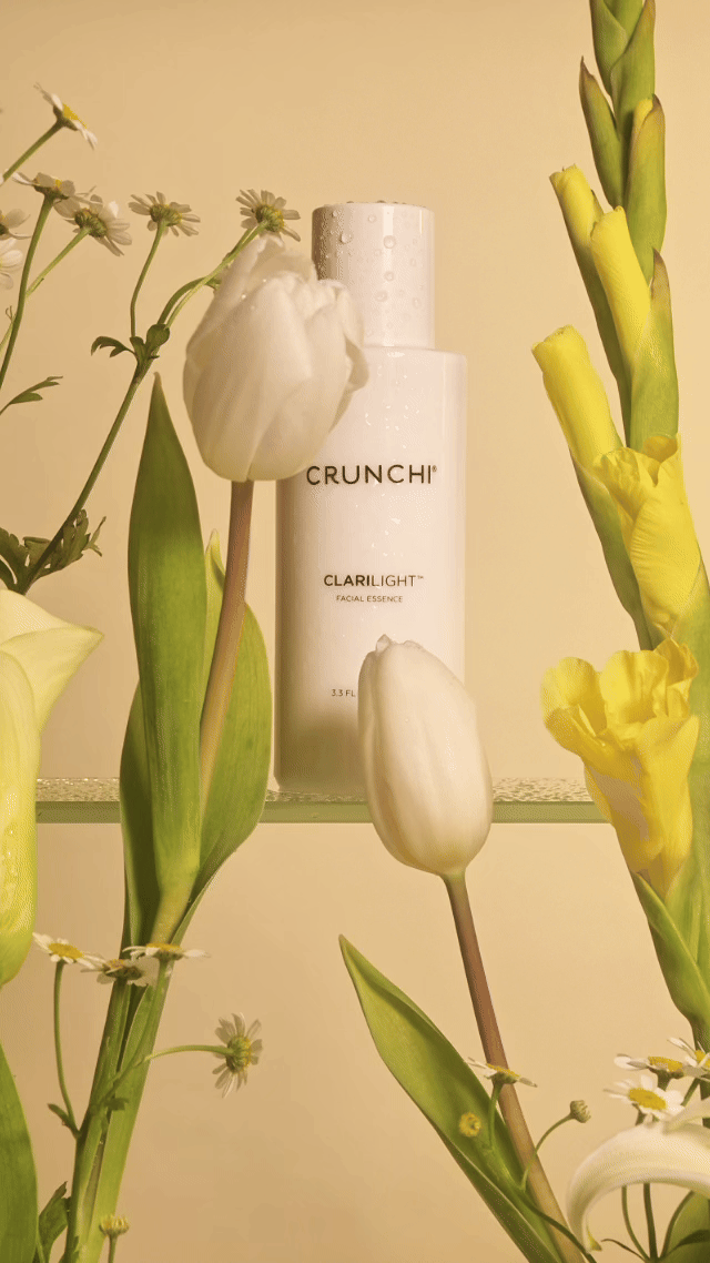 Clean skincare bottle of Crunchi Clarilight facial essence surrounded by white and yellow flowers and green leaves, with a soft butter yellow background.